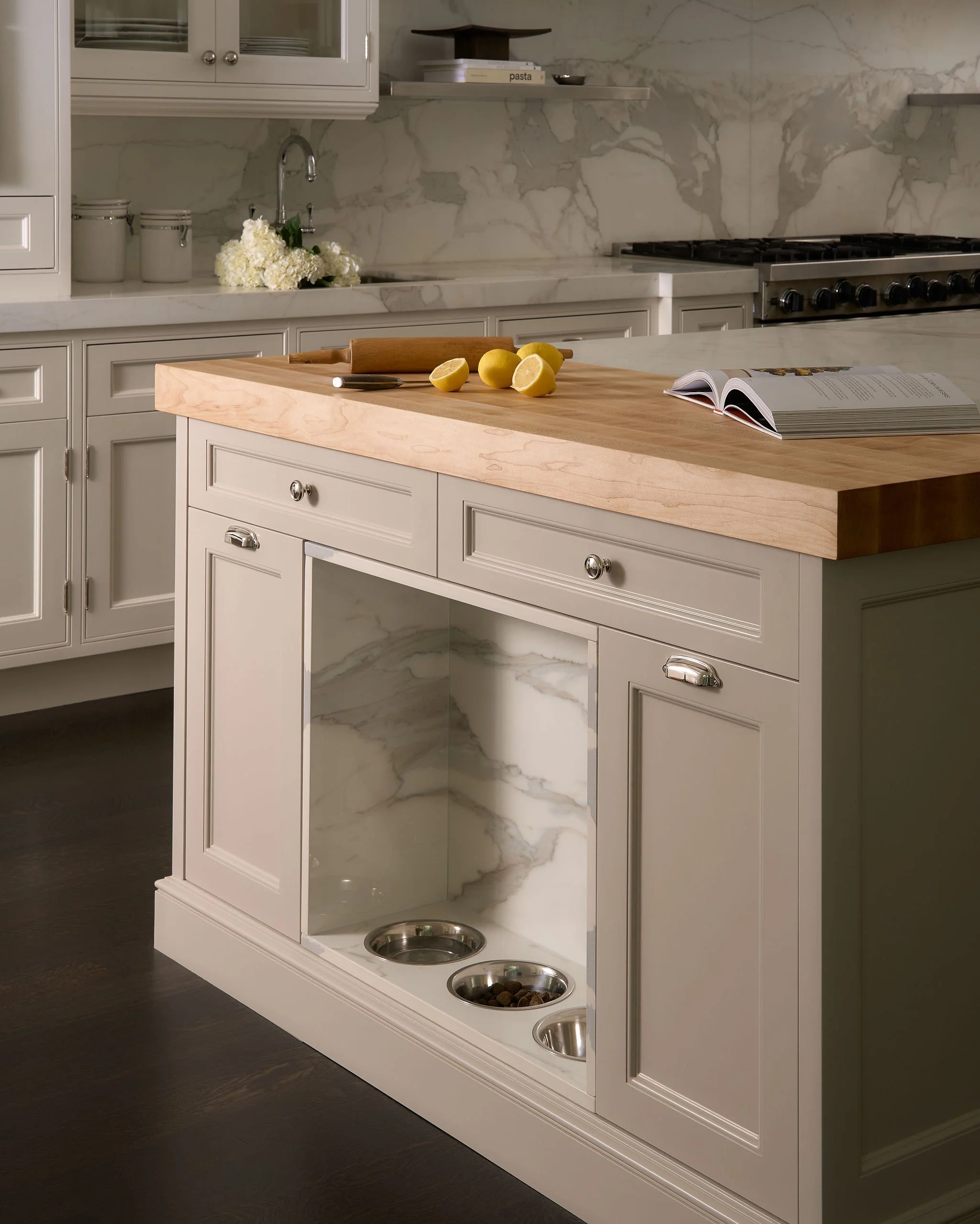 A modern kitchen with a white cabinetry pet feeding station that has four stainless steel bowls, three filled with food and one empty. The station includes a light wood countertop with lemon halves, a book, and a rolling pin. The background features 