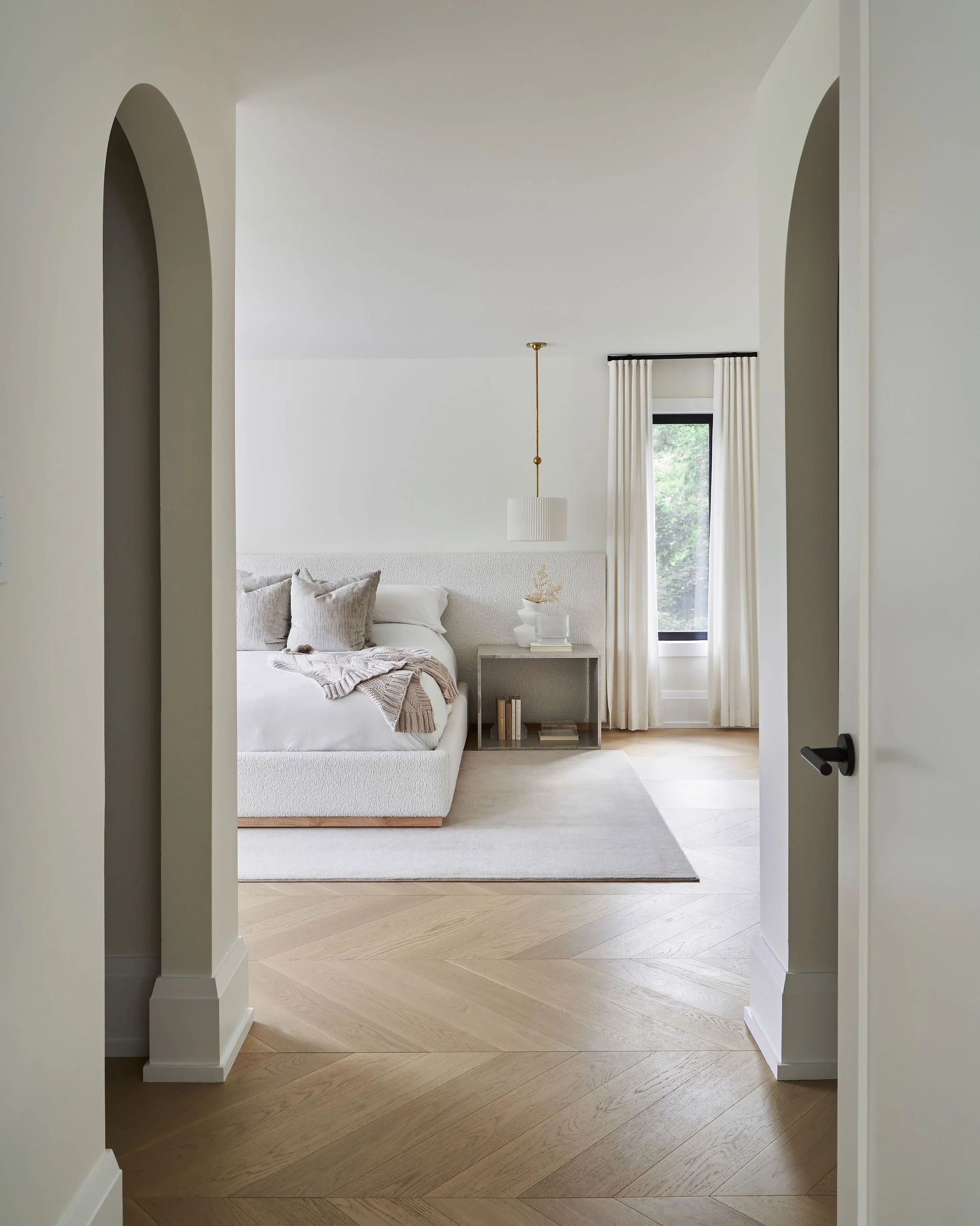 View through an open door into a minimalist bedroom with a white bed, beige pillows, a beige throw, a small side table with a decorative object, a window with white curtains, and a hanging ceiling light.
