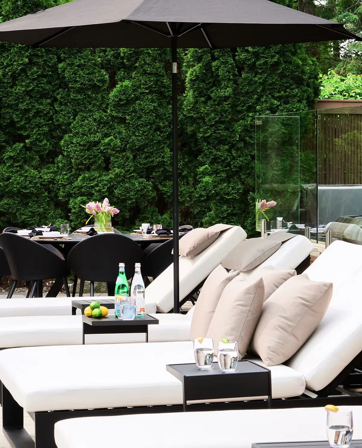 Outdoor patio with white lounge chairs, black side tables, a black dining table set with black chairs, glasses, bottles, and lemons, large umbrellas, and a green hedge in the background.