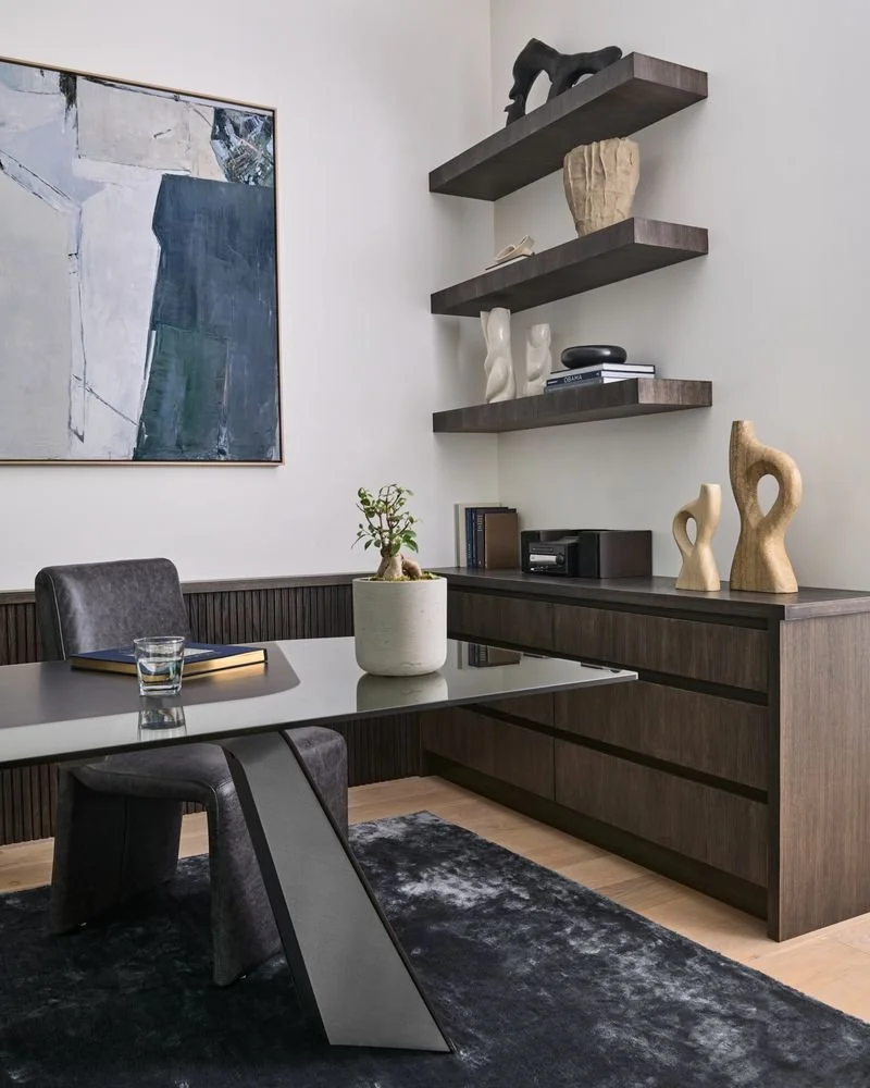 Modern dining room with a black leather chair, a glass of water, a notebook, a potted plant, dark wooden shelves with sculptures and books, a dark wooden sideboard, and a large abstract painting on the wall.