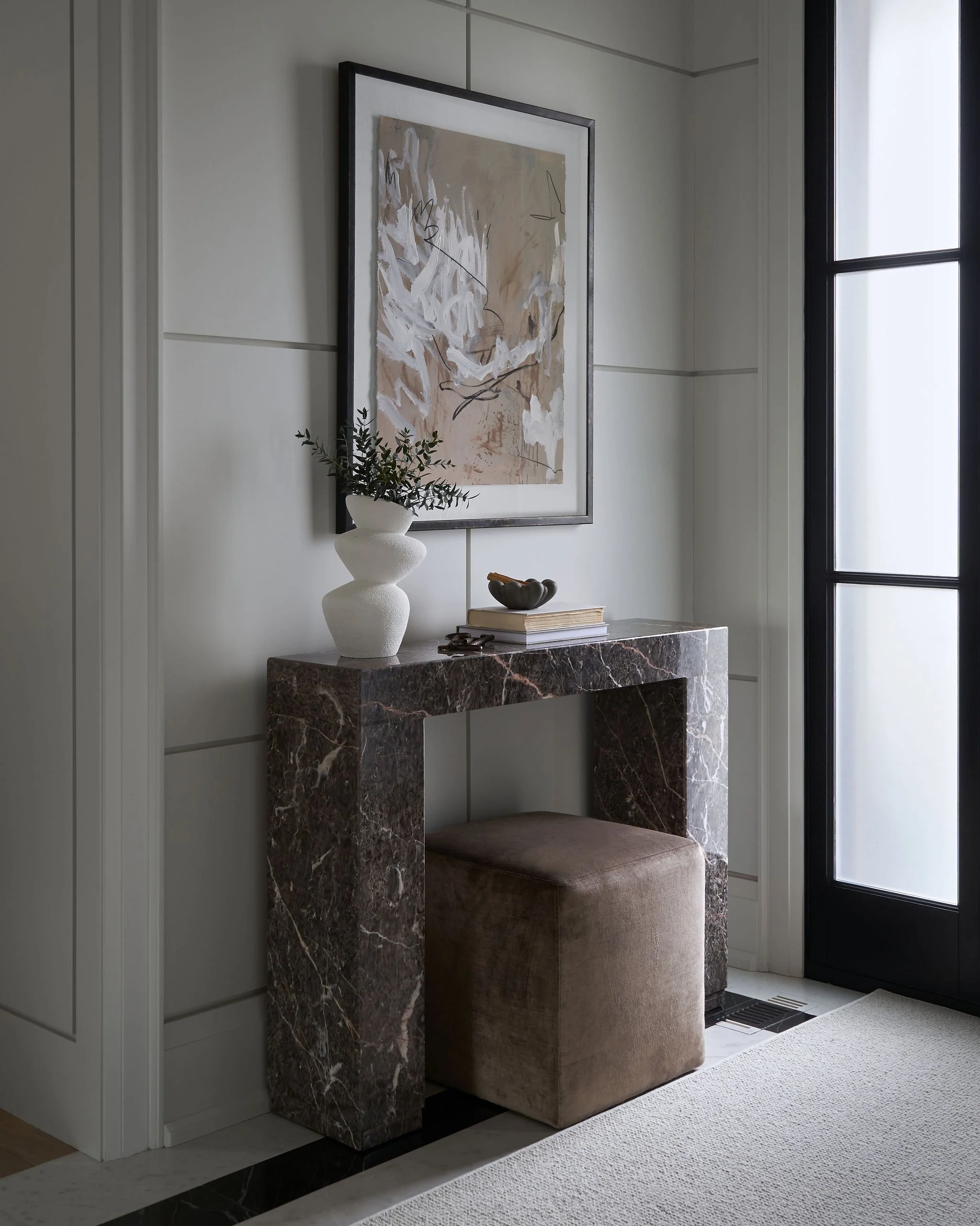 Interior of a modern room with a marble side table, decorative plant, and abstract artwork on the wall near a large window.