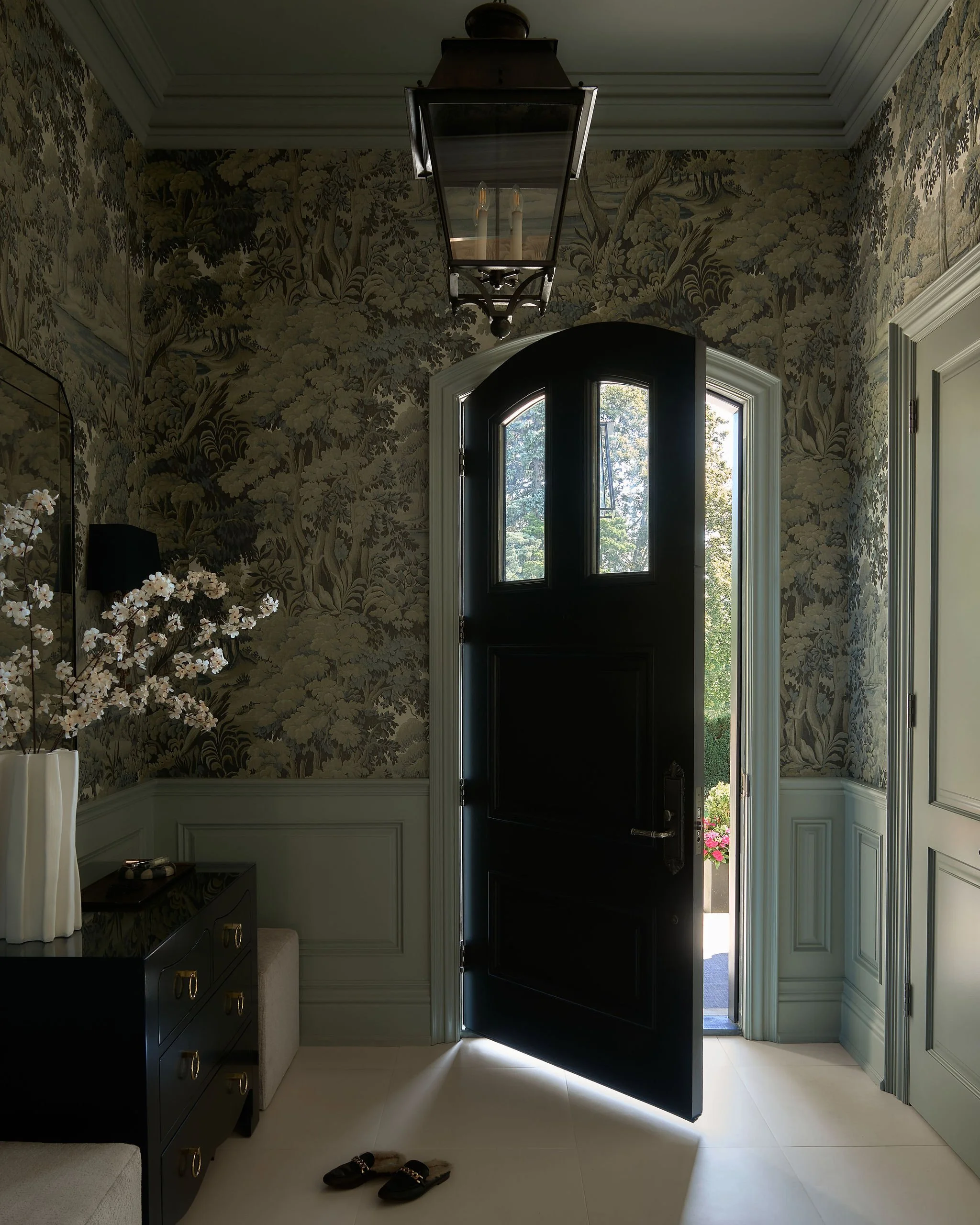 Open black front door leading outside, with trees and flowers visible. Inside, a hallway with wallpaper featuring a floral and tree pattern, white wainscoting, and cream-colored flooring. On the left, a dark dresser with gold handles; on top a white 