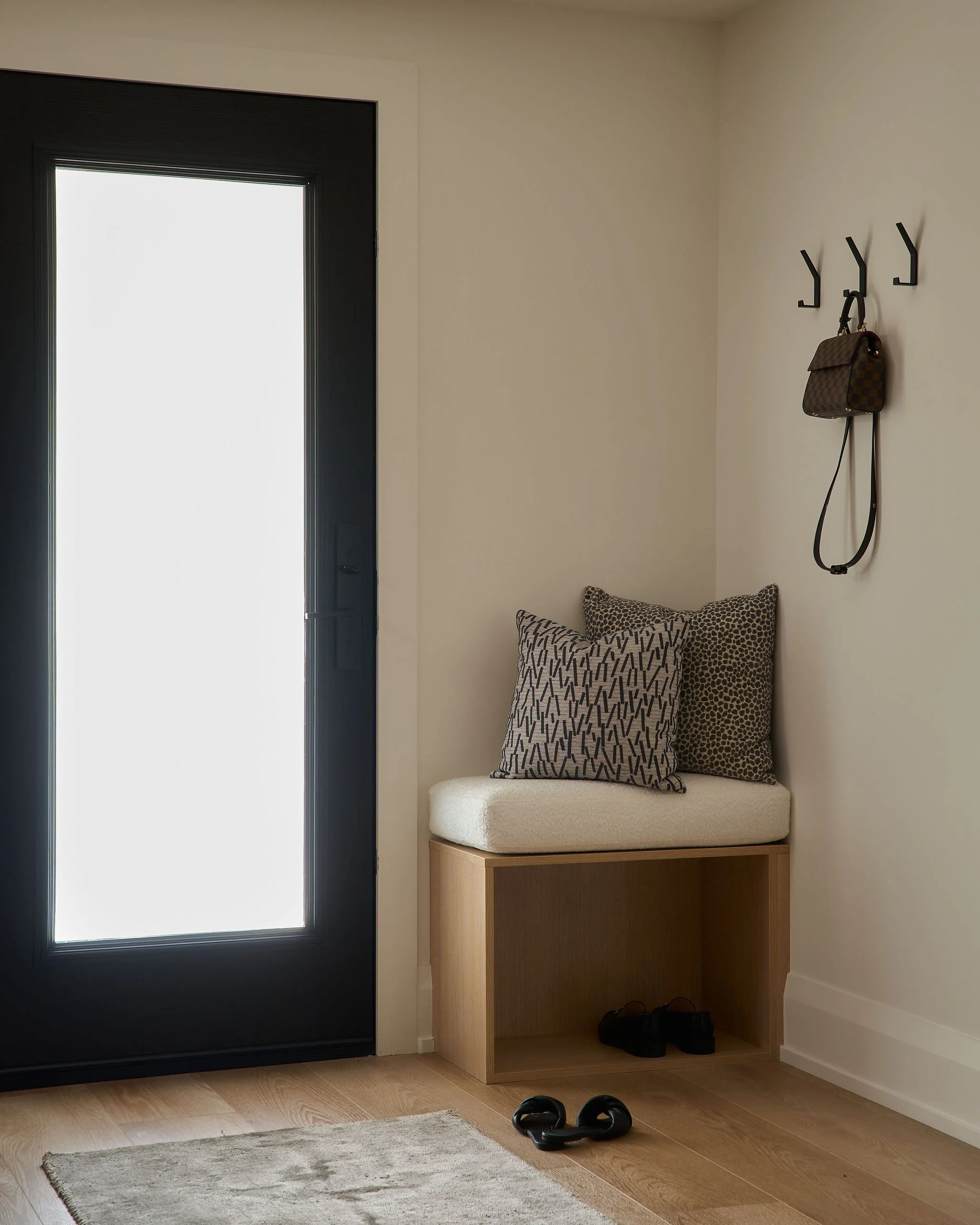 Entryway with black front door, beige walls, a beige cushioned bench, patterned pillows, a small wooden storage unit, black hooks with a small bag hanging, black shoes underneath, black slippers on the floor, and a small rug.