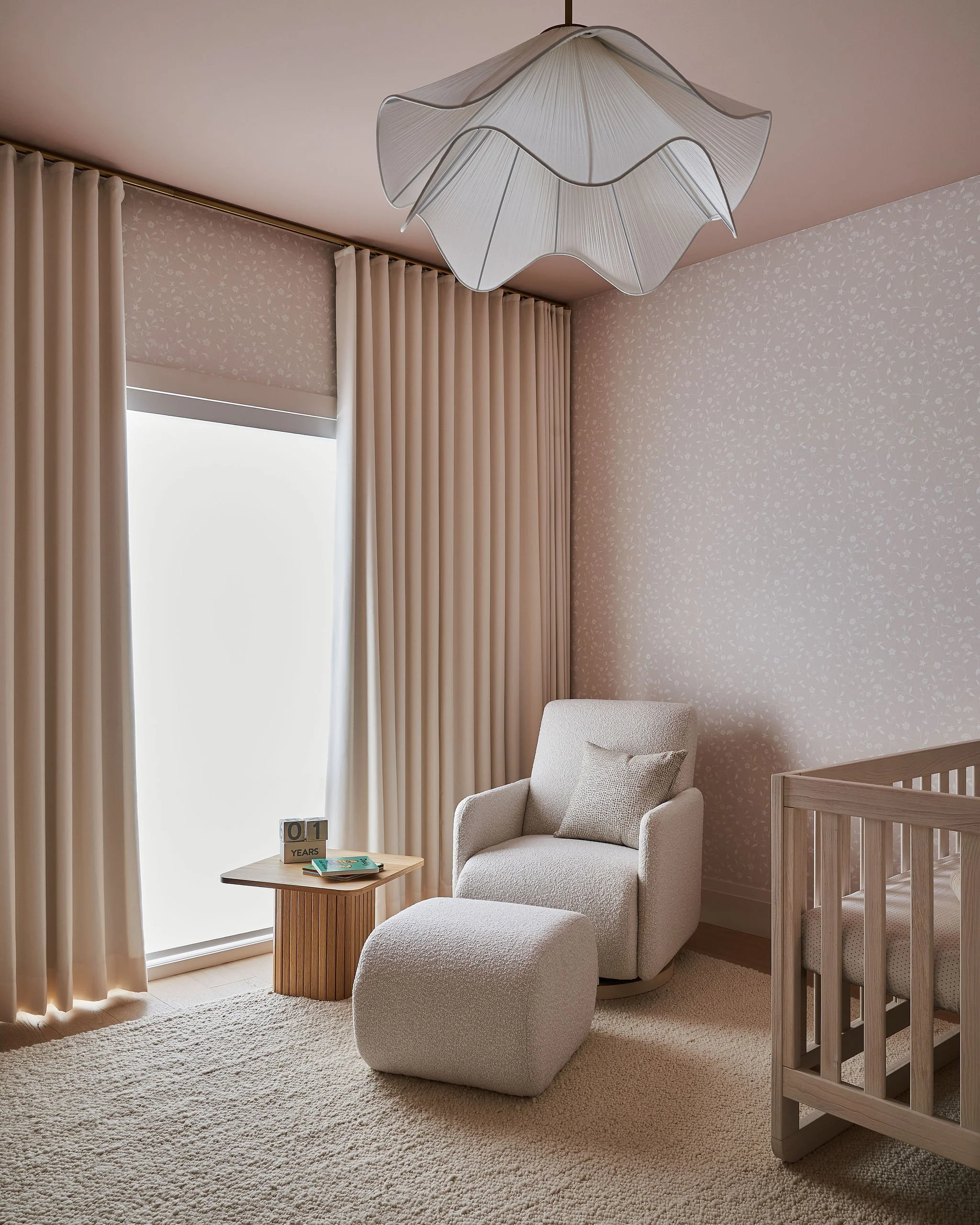 Living room corner with beige armchair, matching ottoman, small wooden side table with books and a sign, large window with cream curtains, modern ceiling light, textured wallpaper, and a wooden crib.