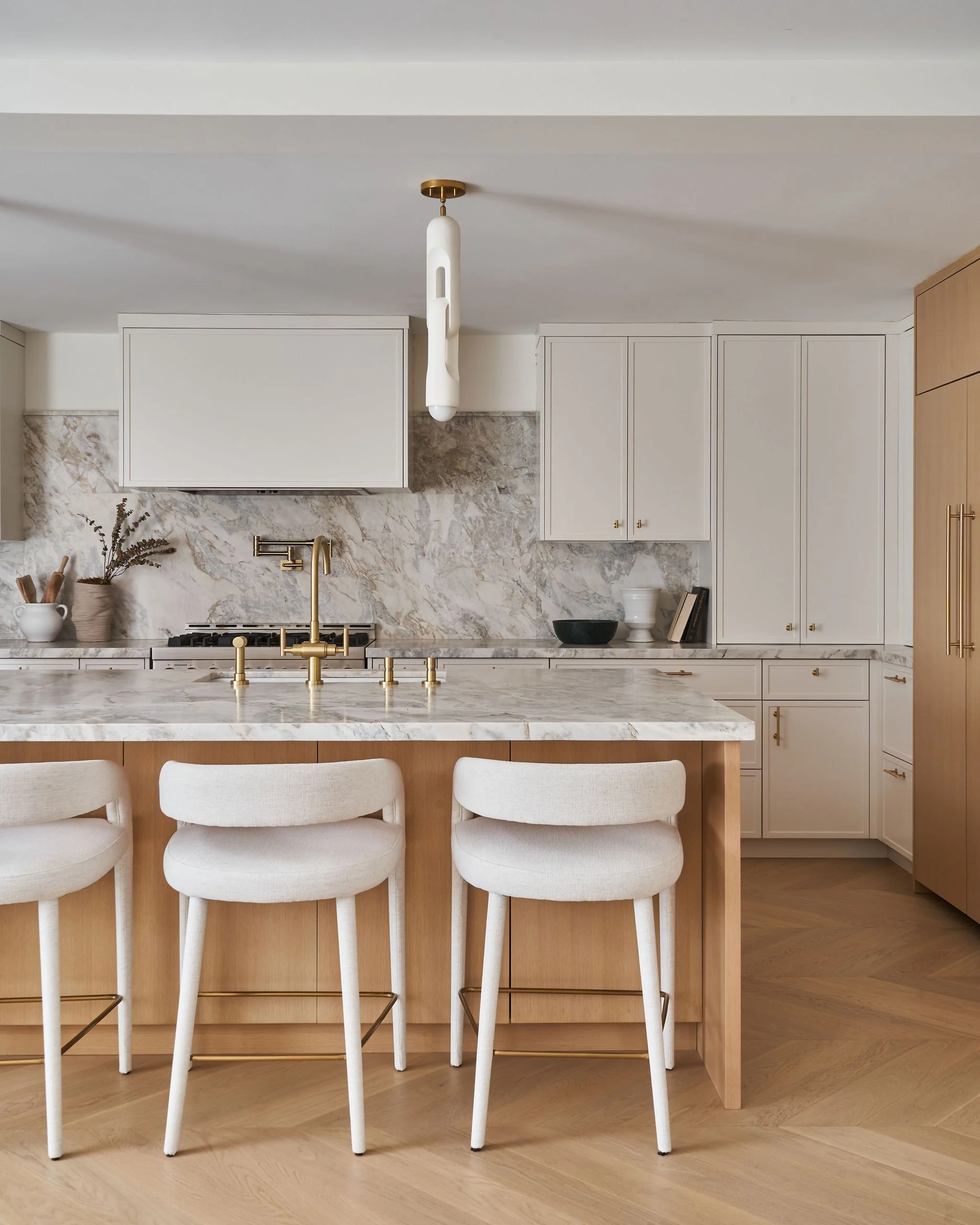 A modern kitchen featuring a marble island with a gold faucet, white cabinetry, and a marble backsplash. There are three white upholstered chairs at the island and a gold pendant light hanging from the ceiling.