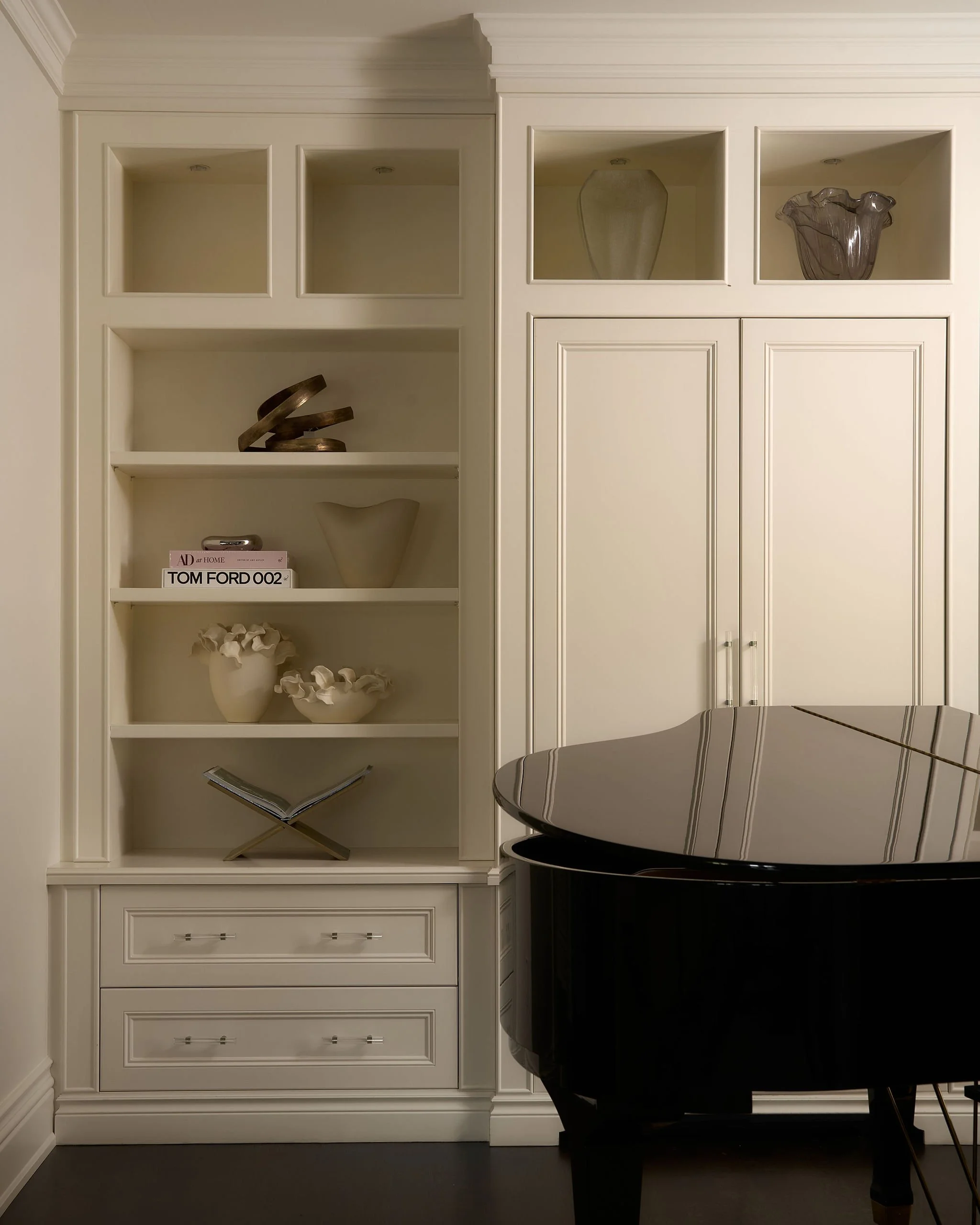 A white built-in bookshelf with multiple compartments and drawers at the bottom, decorated with glass vases, bowls, and books. A shiny black grand piano is partially visible in front of the bookshelf.