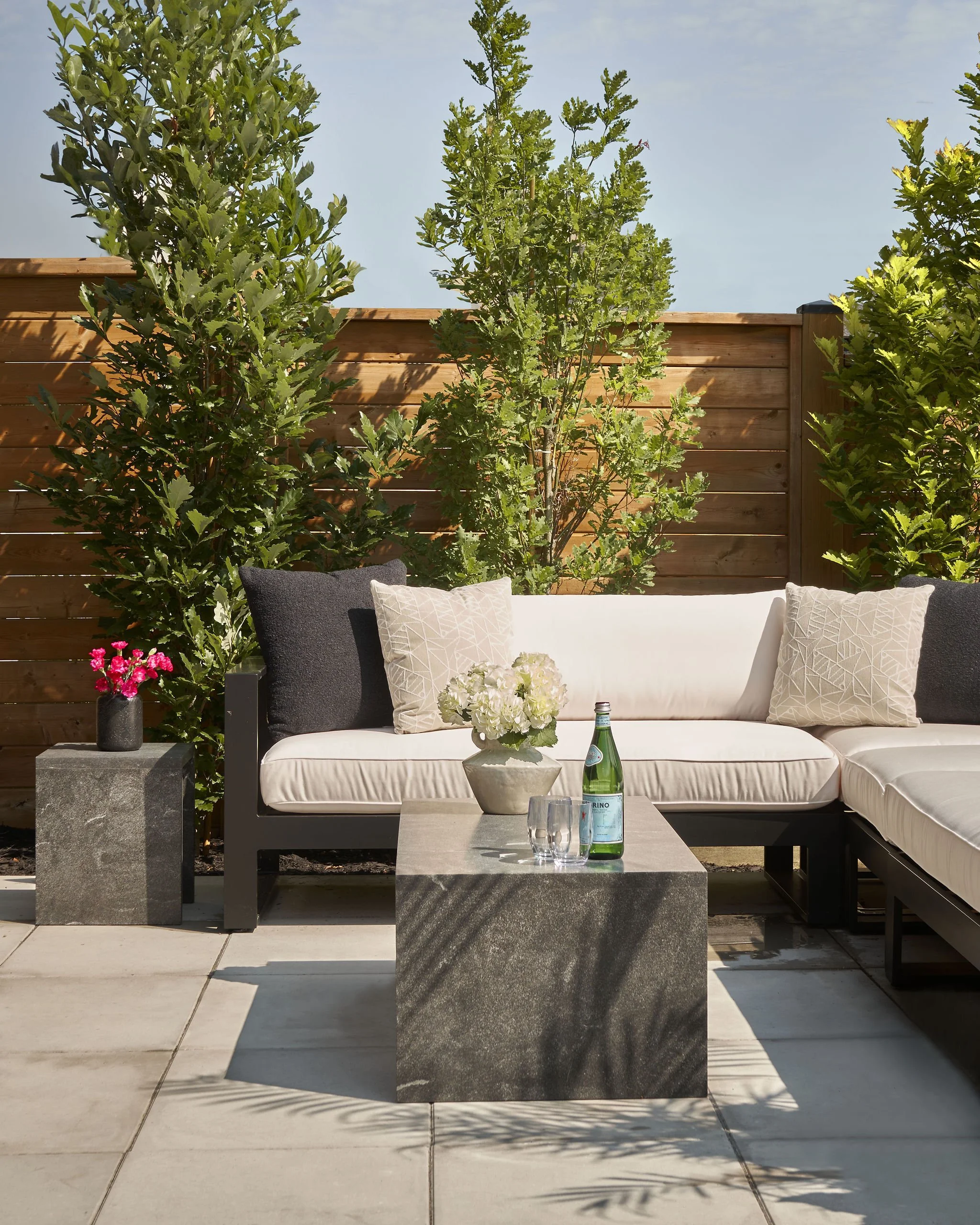 A modern outdoor seating area with a white cushioned sofa, decorative pillows, a black side table holding a vase of pink flowers, a bottle of sparkling water, and glasses. There are tall green plants on either side and a wooden fence in the backgroun