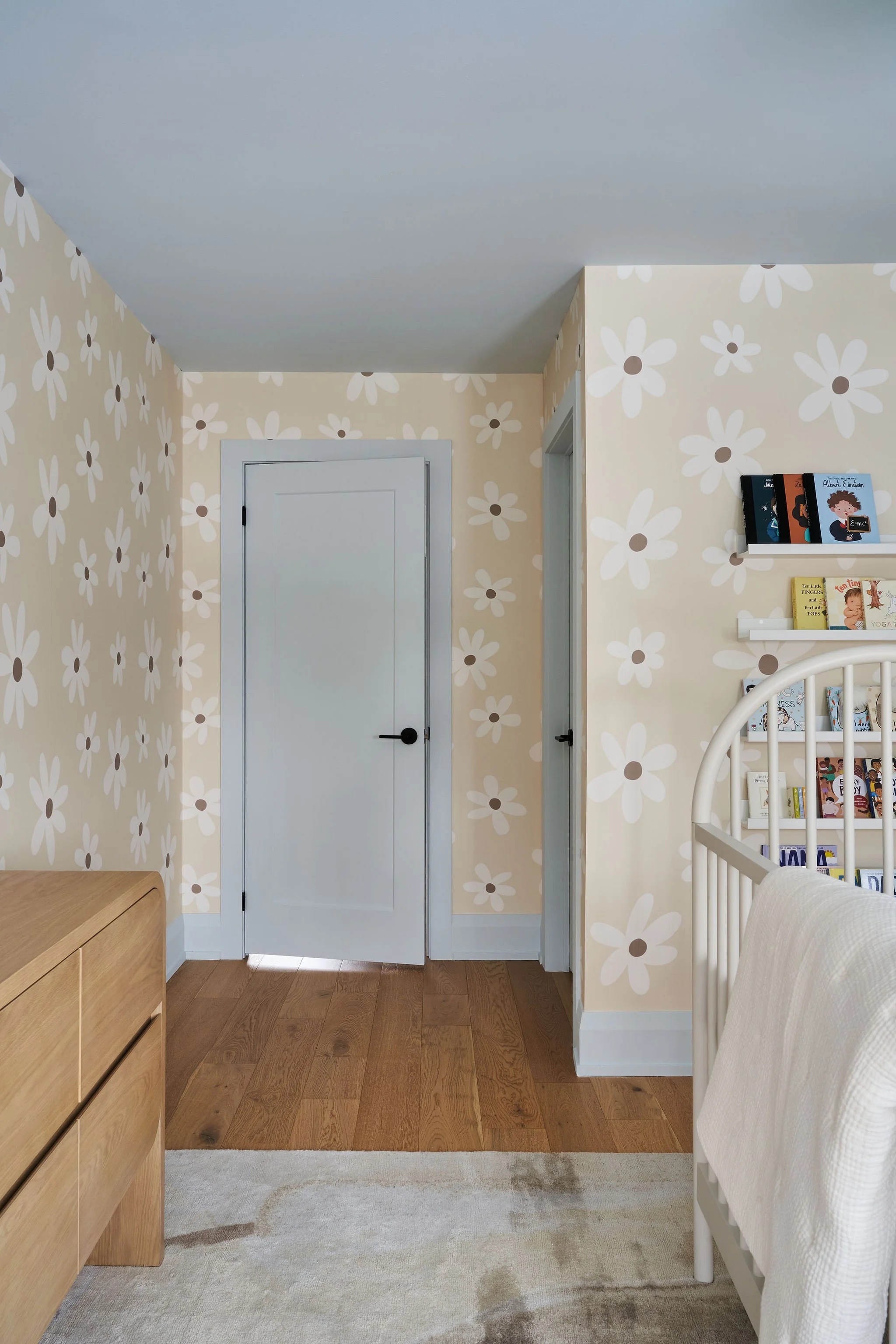 A corner of a bedroom with beige daisy flower wallpaper, white door, bookshelf, wooden dresser, and a beige armchair, with a light-colored rug on the wooden floor.