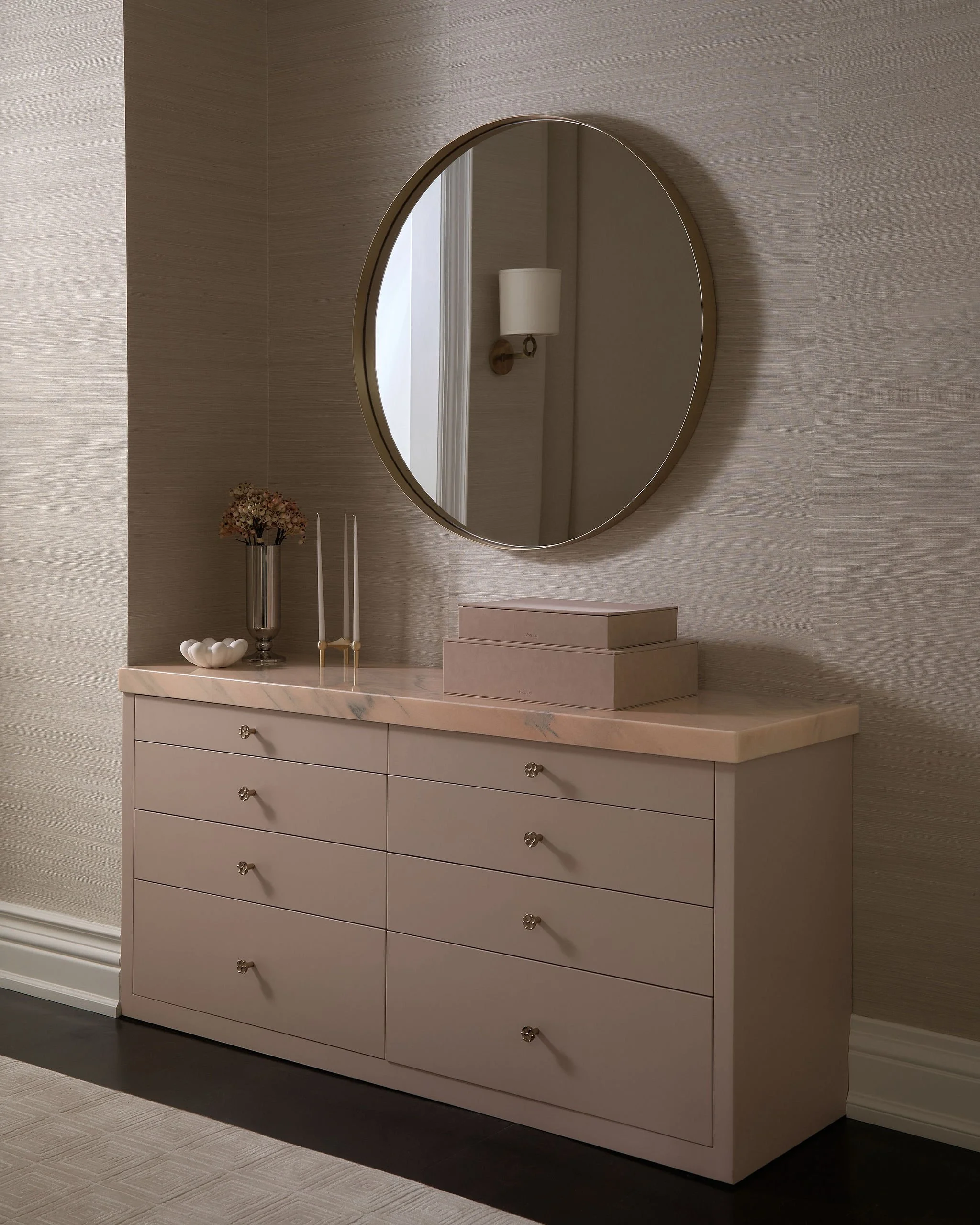 A white dresser with eight drawers and round knobs, topped with a marble surface, is placed against a beige, textured wall. Above the dresser, a large round mirror reflects a wall-mounted light fixture and part of a doorway. On the left side of the d