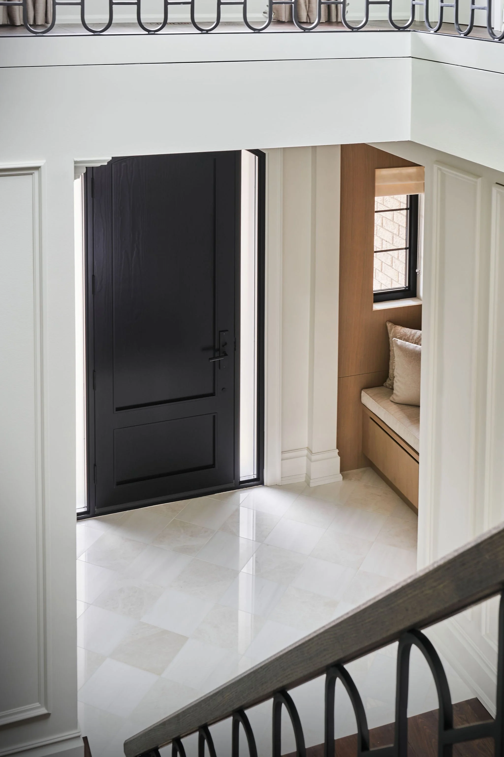 View of a black front door with glass panels next to a wall with a built-in window seat, decorated with a pillow and a window with a shade, in a brightly lit interior space.