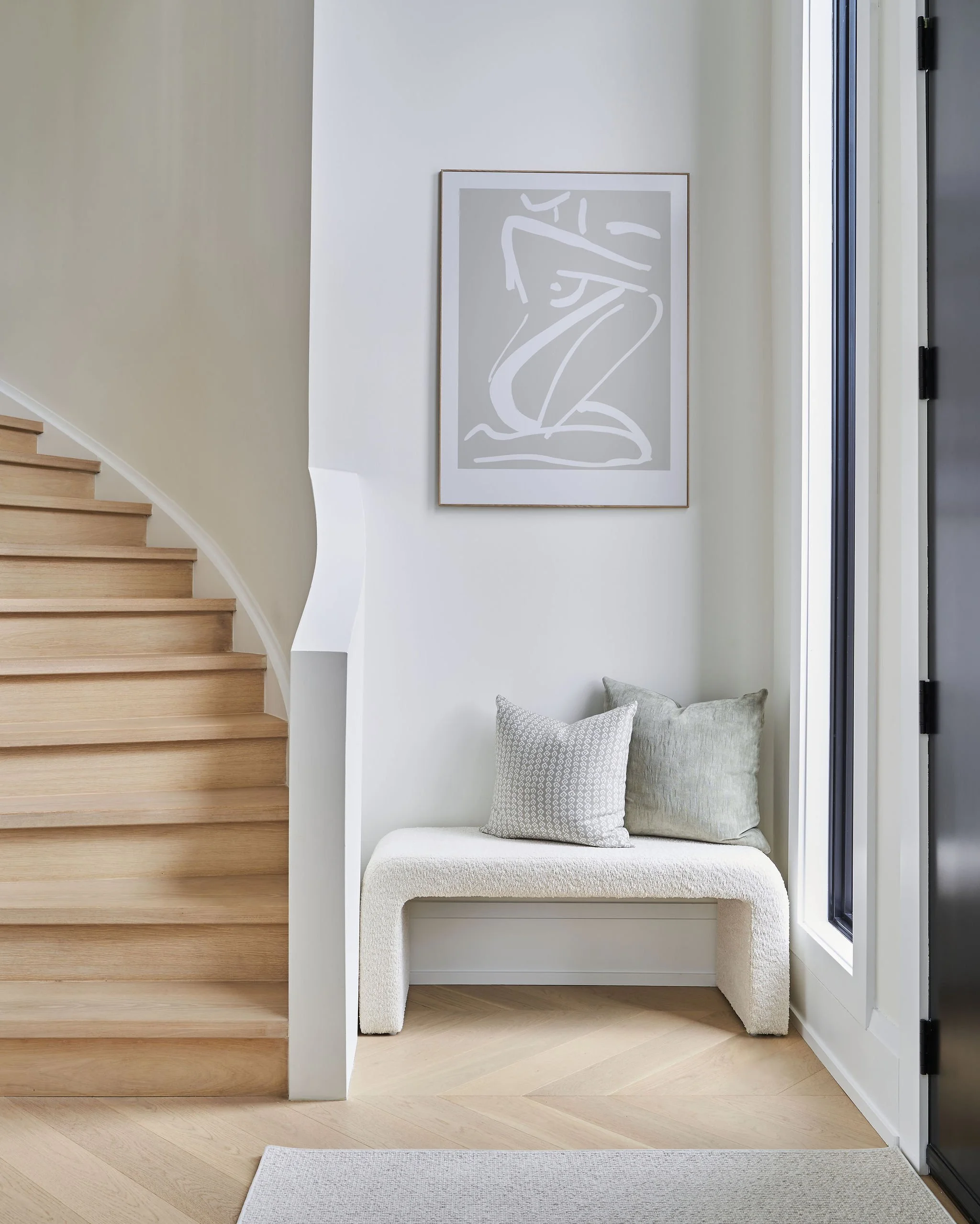 Interior of a modern home with light wood steps on the left, a white wall with a minimalist line art painting in a wooden frame, a small cushioned seating area with two pillows, and a glass door on the right allowing natural light.
