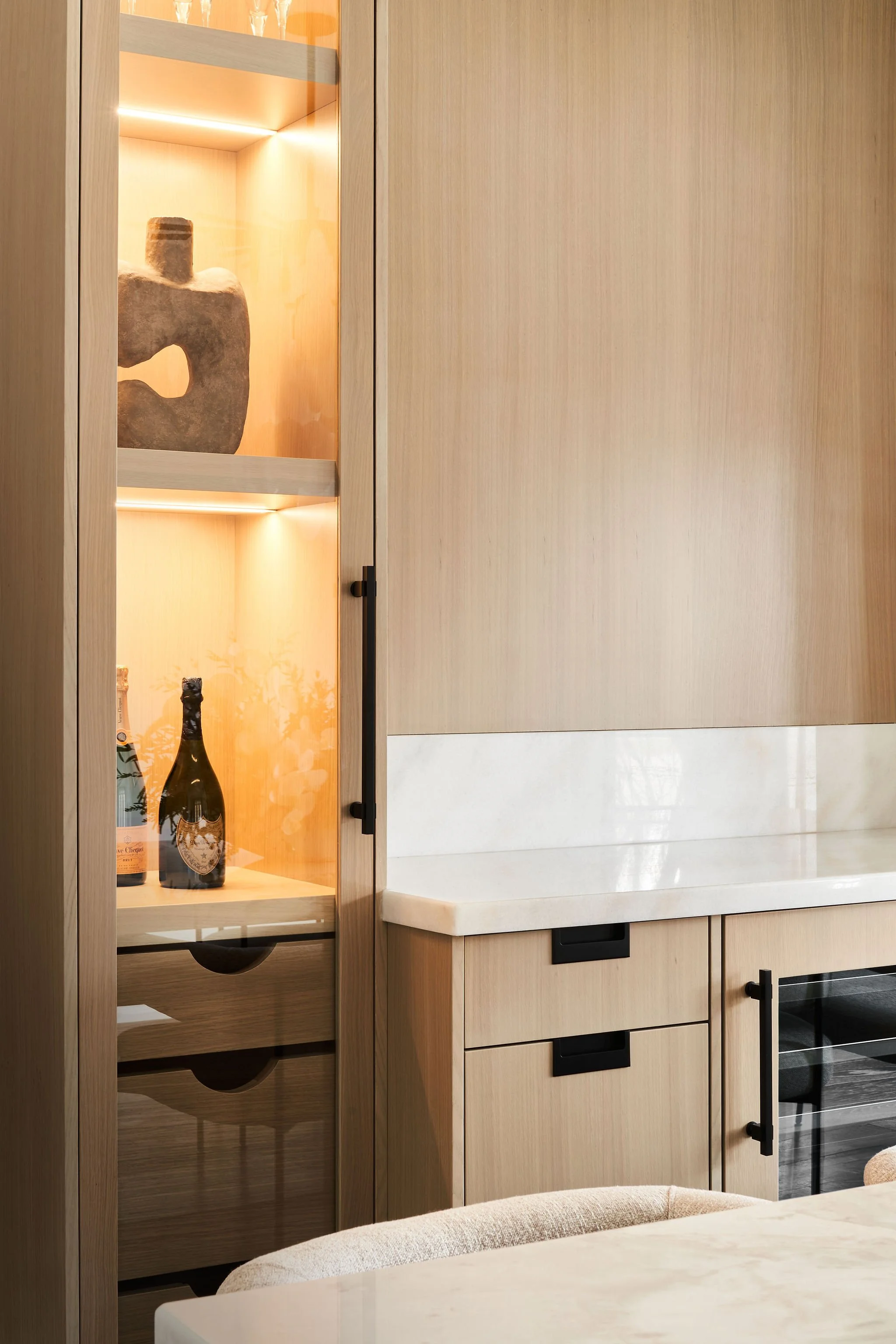 A corner of a wooden cabinet with a glass door displaying bottles of champagne and a decorative stone sculpture inside, situated next to a beige marble countertop.