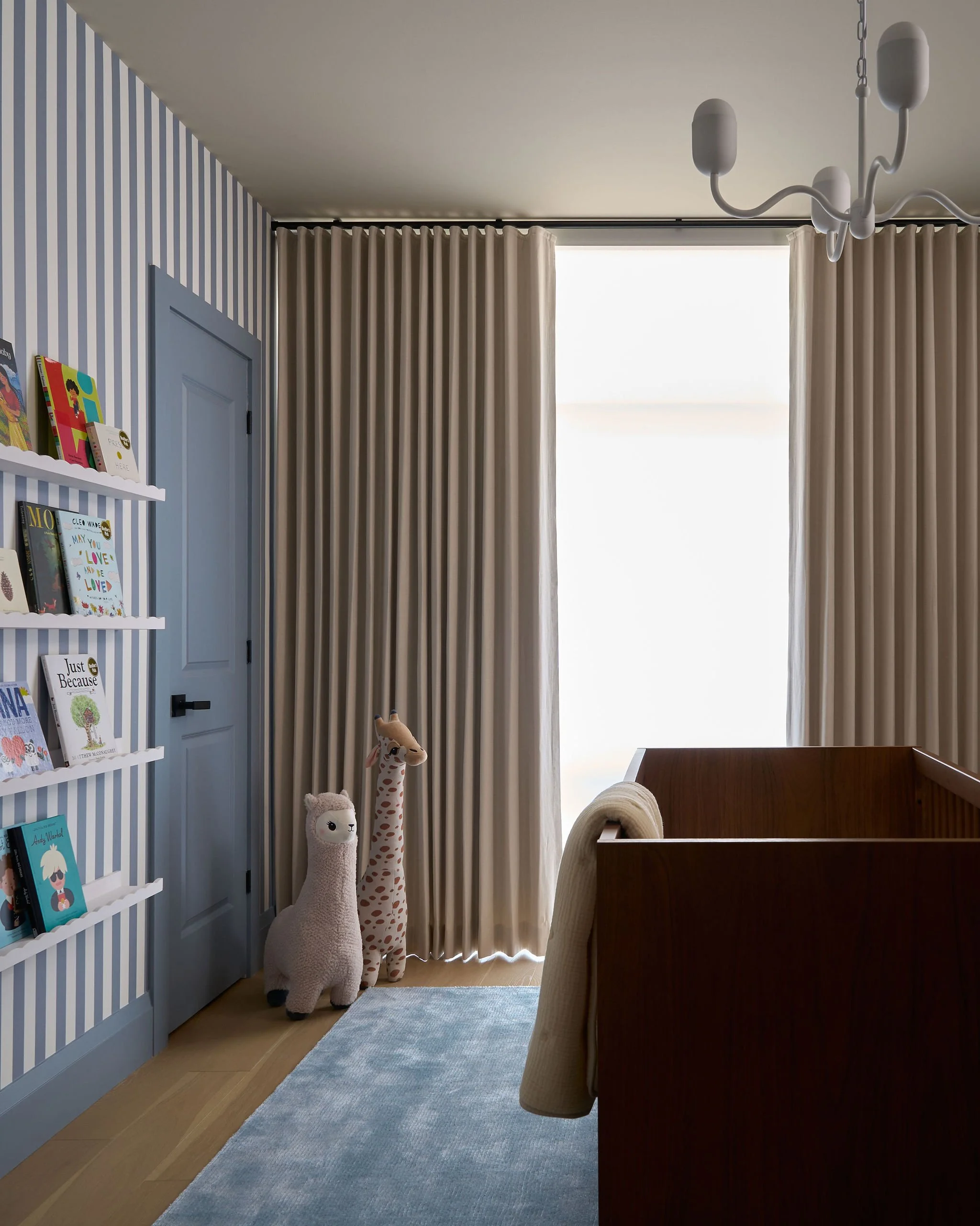 A cozy nursery room with striped wallpaper, books on a white bookshelf, plush animal toys, beige curtains, and a glimpse of a wooden crib.