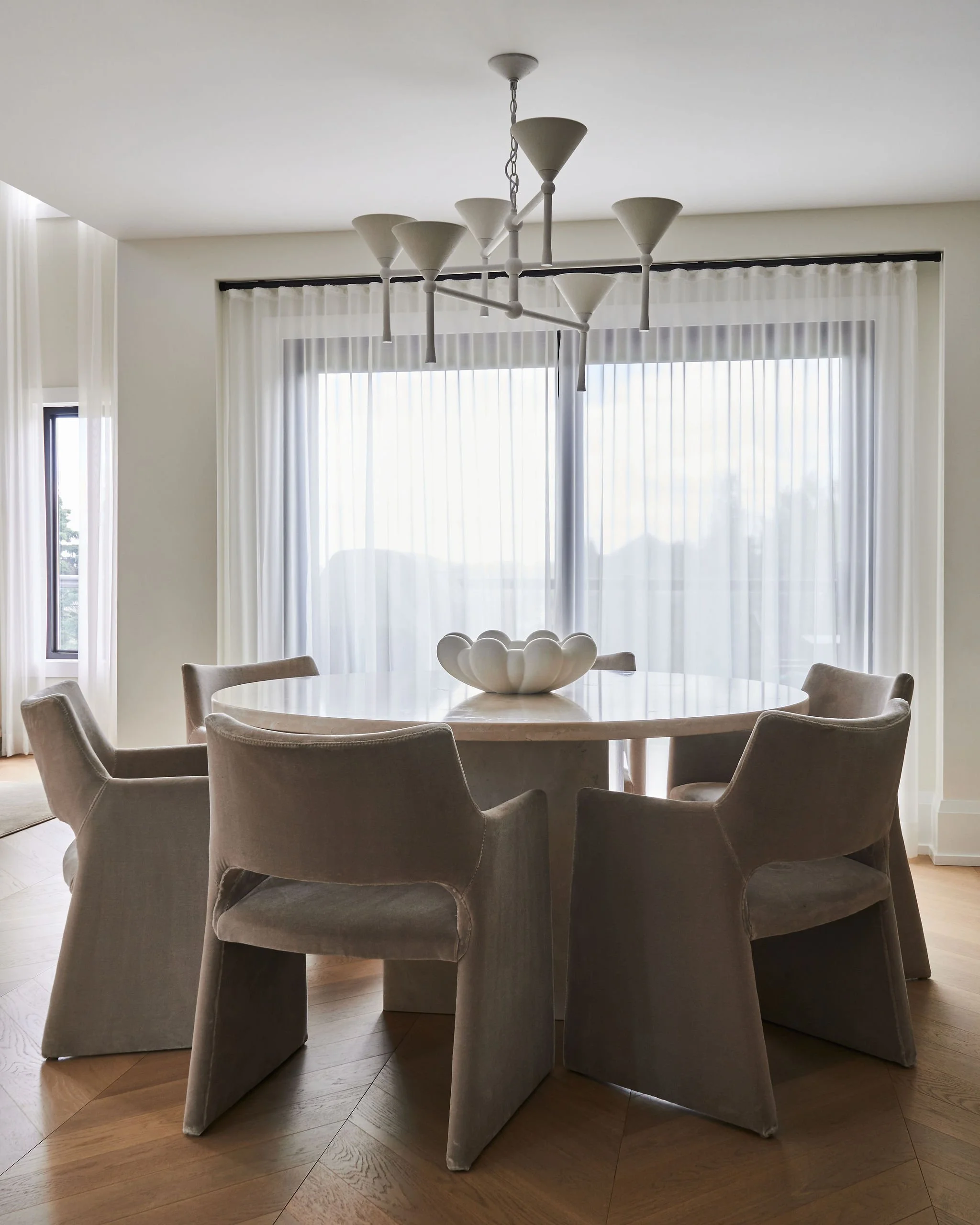 A round dining table surrounded by eight upholstered armchairs in a modern, well-lit room with floor-to-ceiling curtains and a contemporary chandelier hanging above.