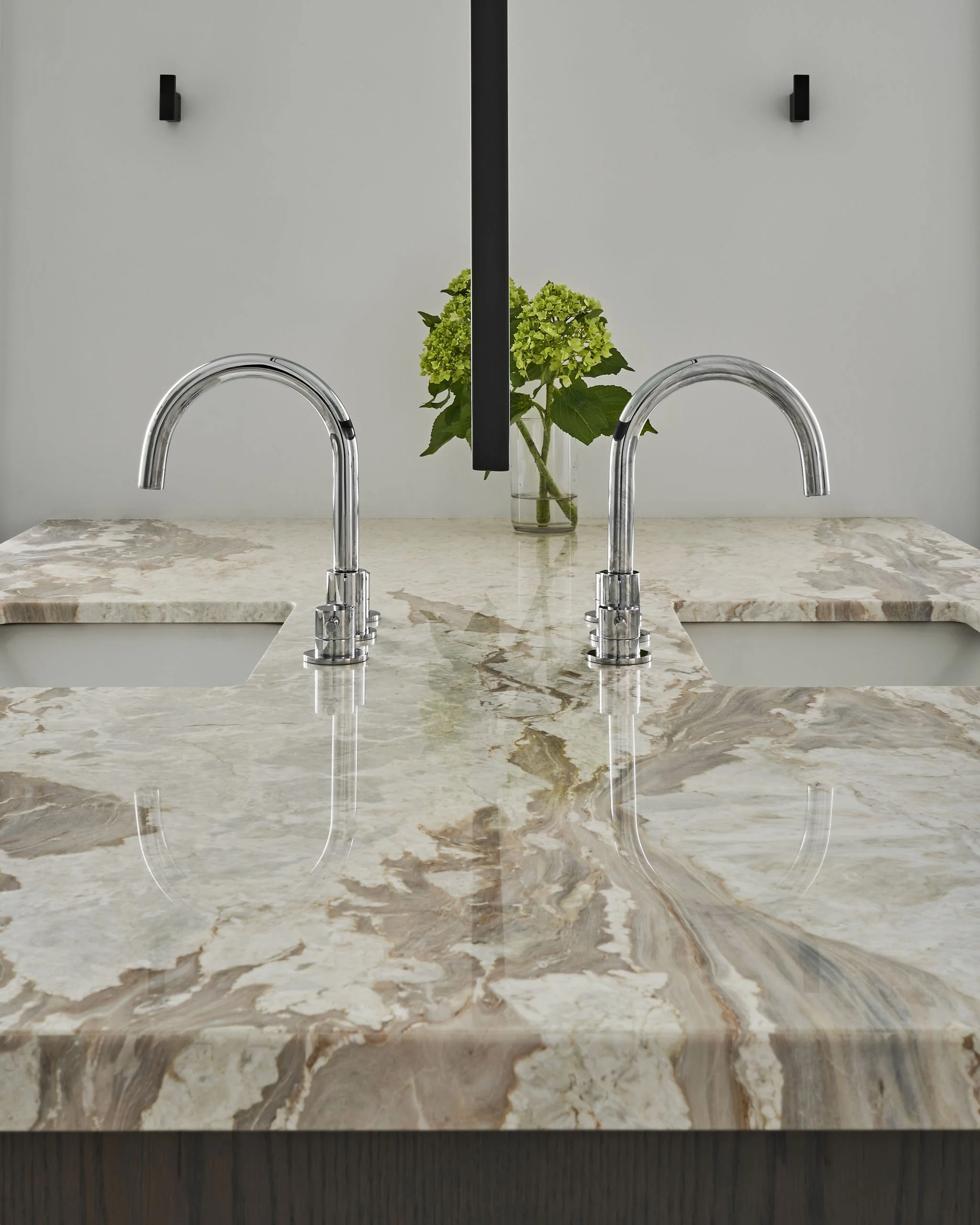 A marble kitchen countertop with two silver kitchen faucets and a glass vase with green hydrangea flowers in the background.