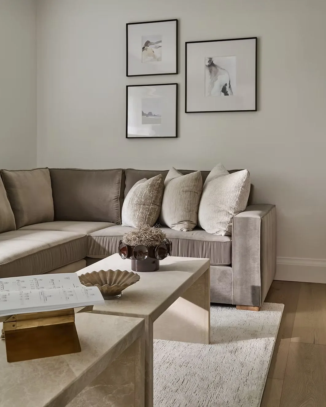 Living room with beige sectional sofa, light-colored pillows, framed minimalistic art on white wall, white coffee table with decorative items, and light wood flooring.