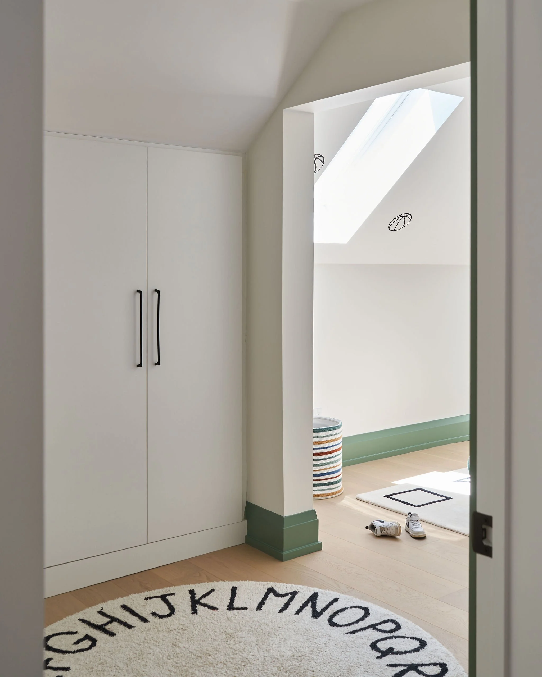 Entryway with a round rug featuring the alphabet, a pair of white sneakers on the floor, a coat closet with black handles, and a view into another room with a striped laundry basket, a small rug, and a shoe.