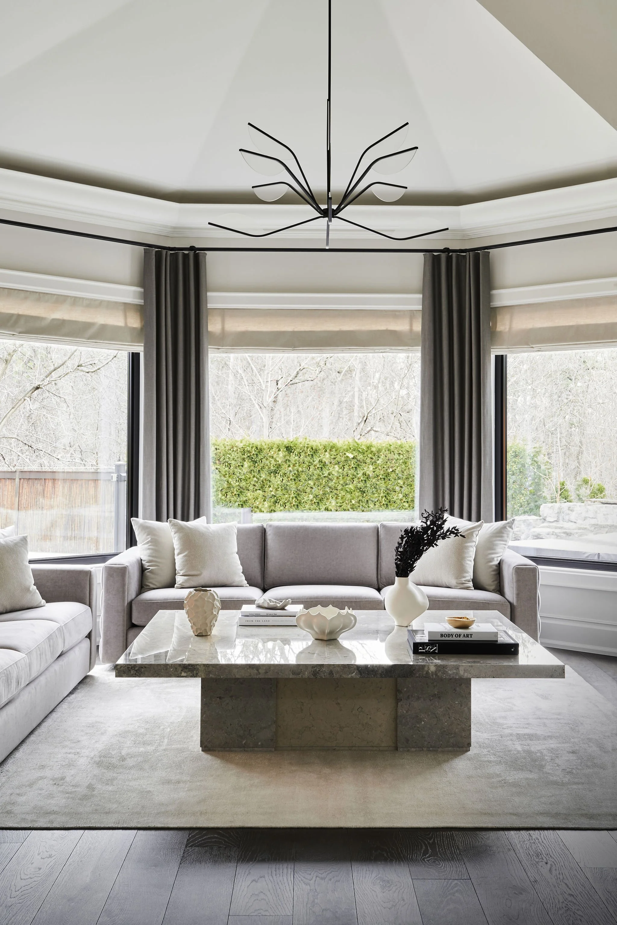 Modern living room with large windows, gray curtains, a gray sectional sofa with pillows, and a marble coffee table with decorative vases and books. There is a contemporary black metal chandelier hanging from the ceiling.