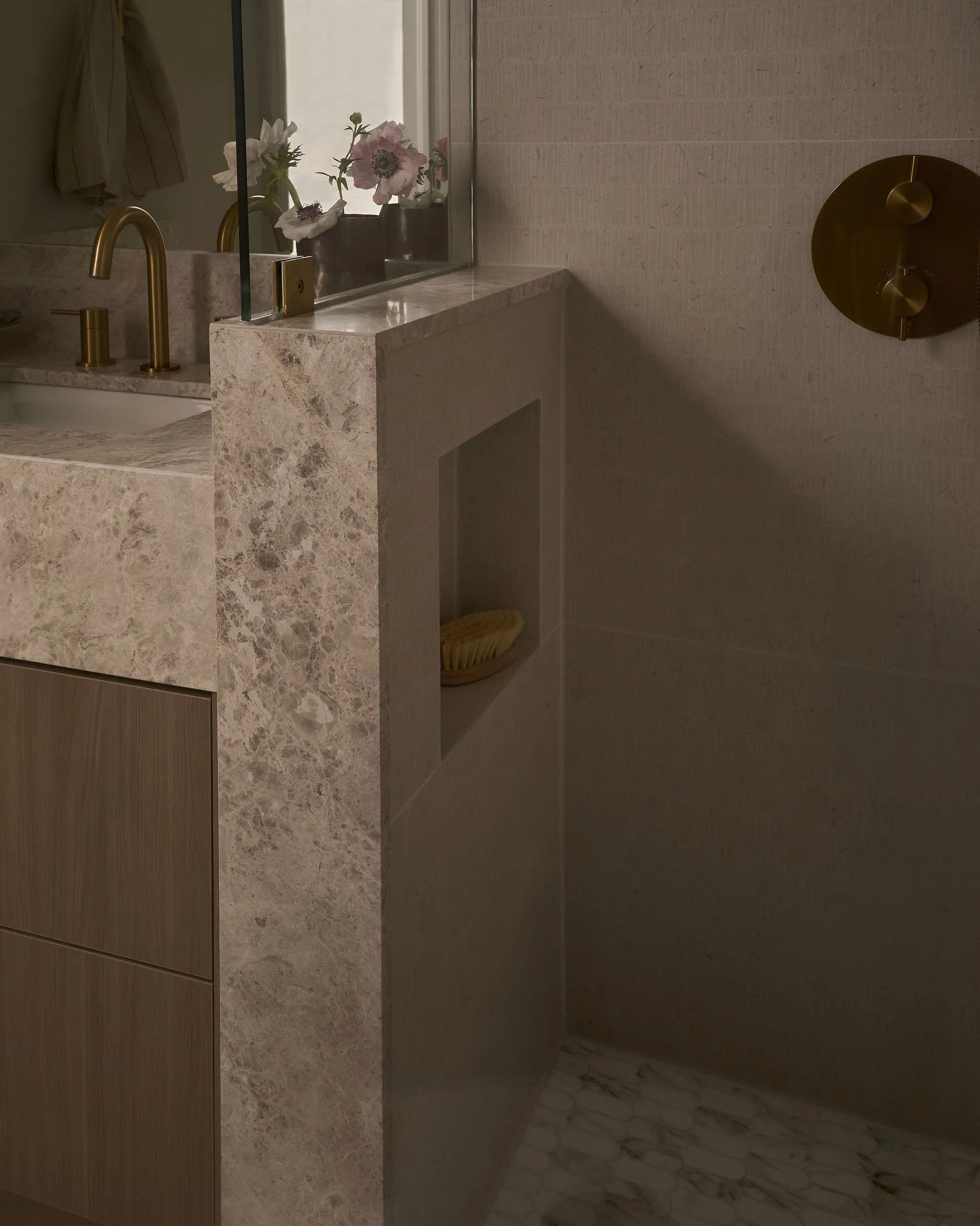 A bathroom vanity with a marble countertop, a gold faucet, pink flowers in a black vase, a mirror, and beige walls