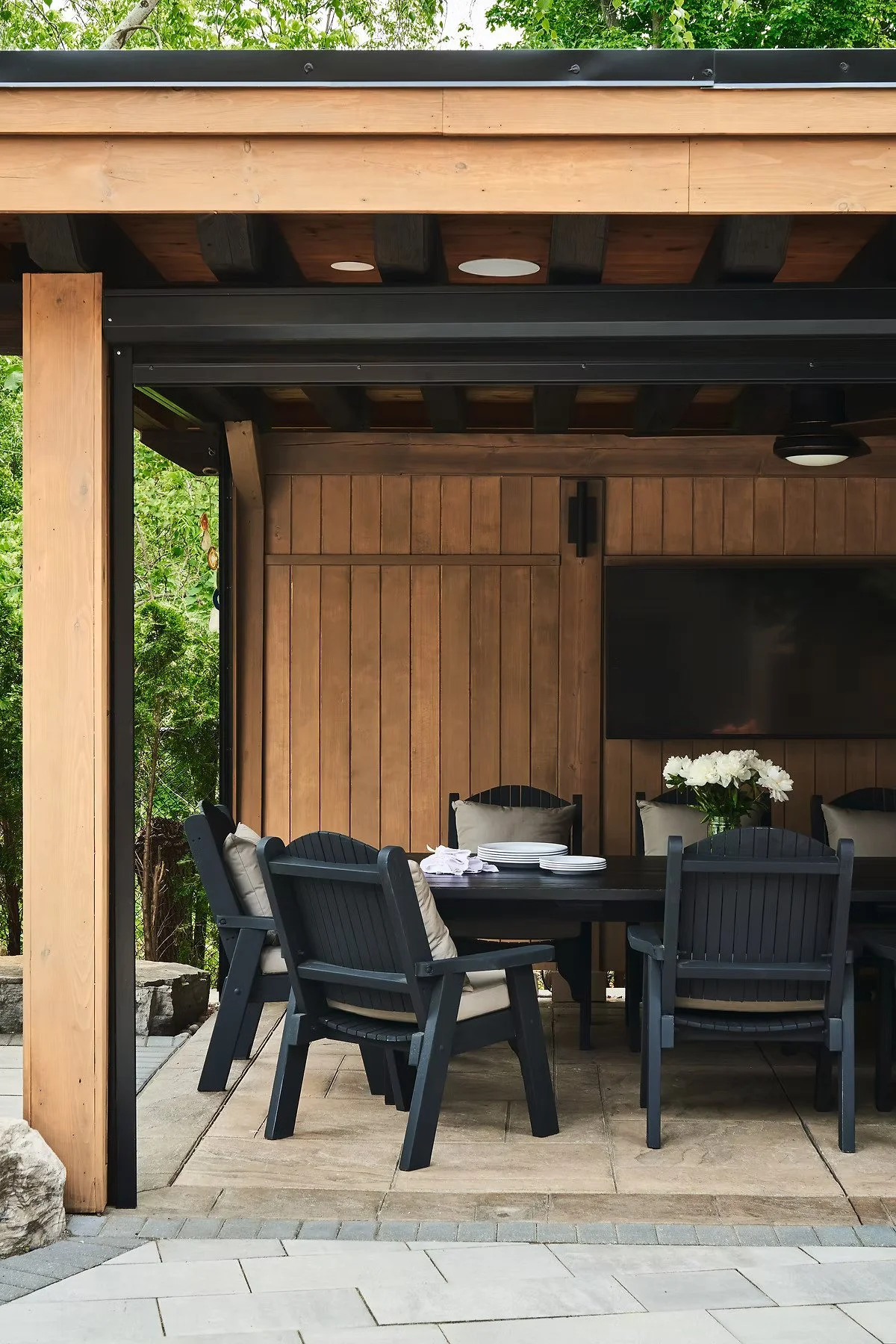 A covered outdoor dining area with a wooden wall and modern black outdoor furniture. There is a dining table with white plates, napkins, and a vase of white flowers. The ceiling is wooden with black beams and a mounted black television. Green foliage