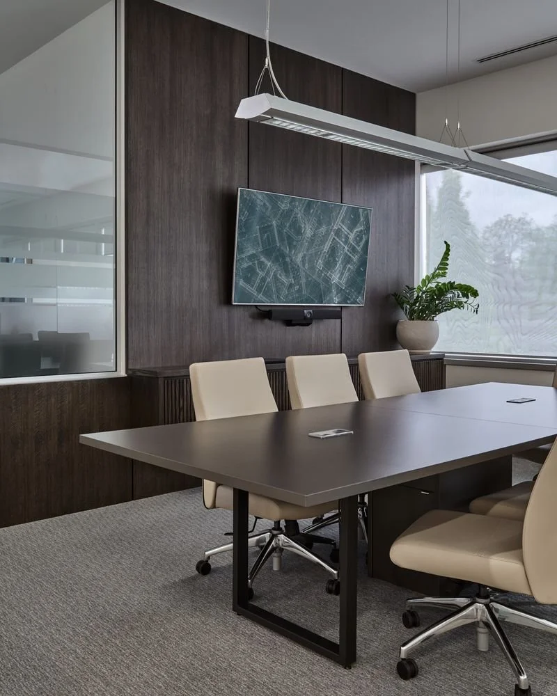 Empty conference room with a large black table, four beige chairs, a wall-mounted screen displaying a map or blueprint, a potted plant, and large windows with blinds.