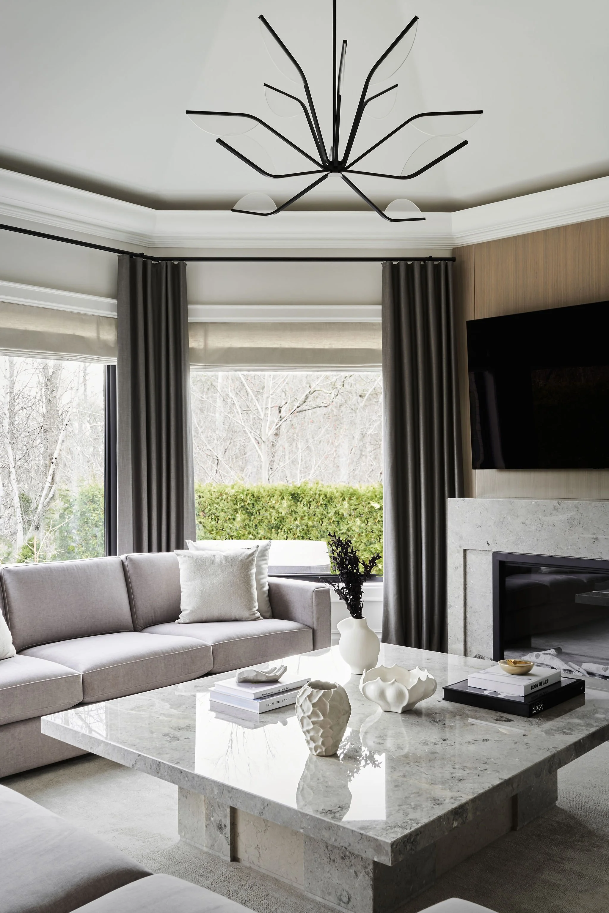 Modern living room with gray sofa, white cushions, marble coffee table with white decorative vases, and gray curtains. Large window showing trees outside, ceiling with black and glass light fixture, and a wall-mounted TV beside a fireplace.