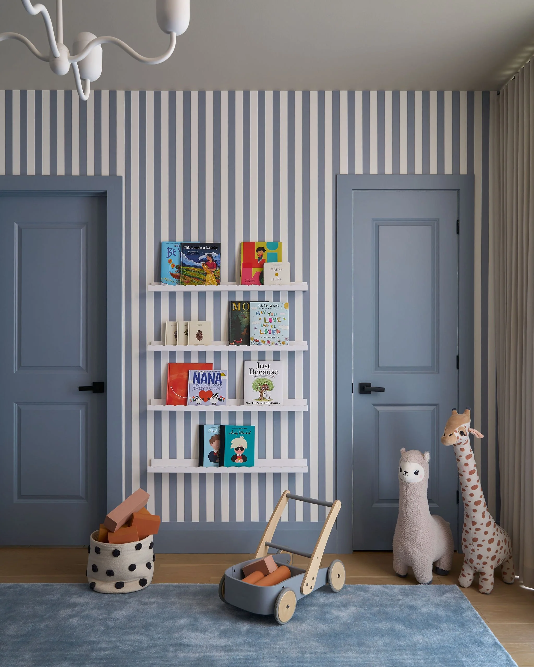 Children's playroom with striped wallpaper, a white bookshelf holding children's books, stuffed animals resembling a llama and a giraffe, a toy cart filled with foam blocks, and a fabric storage bin with foam blocks. The room features a blue door and