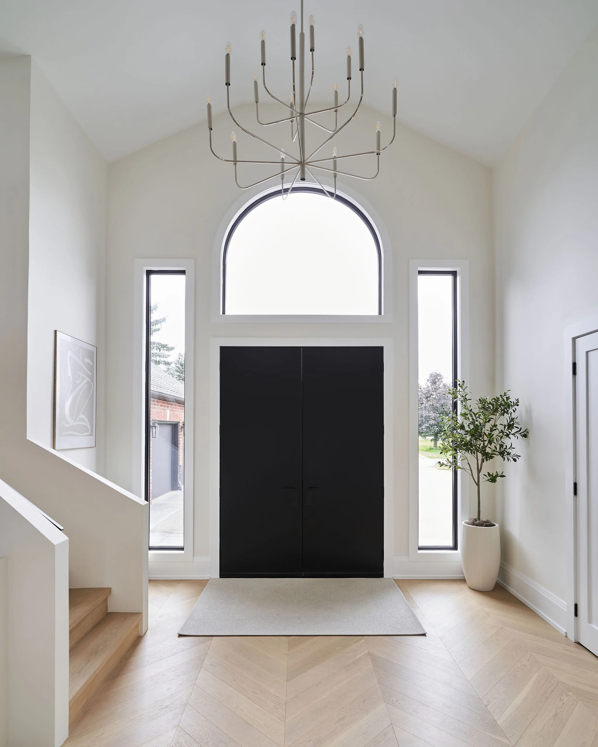 Entryway with black cabinet, tall arched window, and two smaller windows, a chandelier, a potted plant, and a light-colored rug on wooden floor.