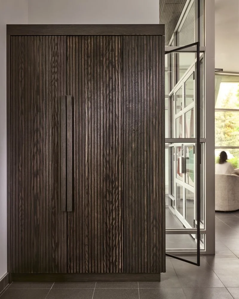 Wooden cabinet with vertical grooves and a long handle, next to open glass doors leading outside to a sitting area.