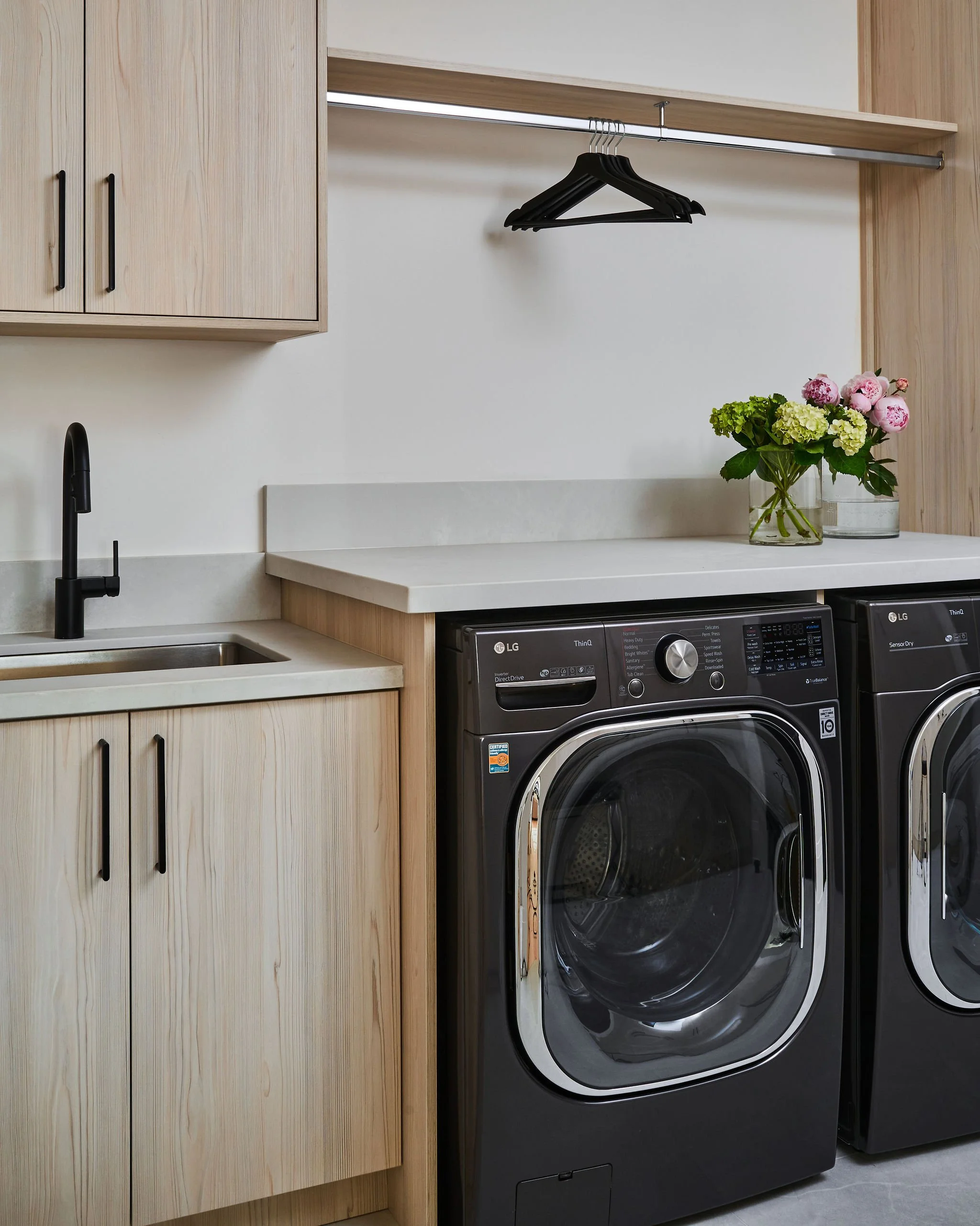 A laundry room with wooden cabinets, a white countertop, a black faucet, and a black front-loading washing machine next to a black dryer. There are two glass vases with pink and green flowers on the countertop.