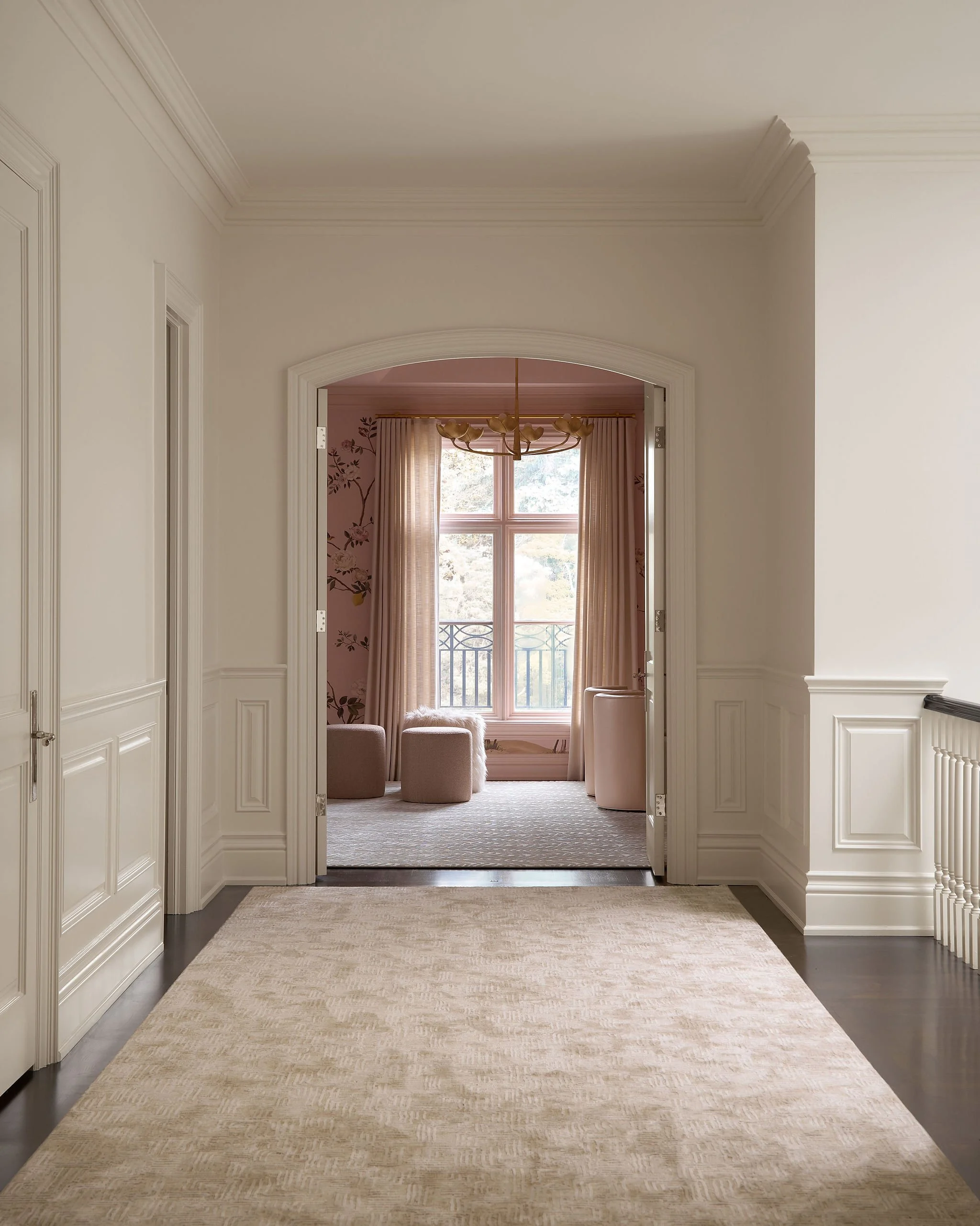 View of an elegant hallway leading to a sitting area with large window, pink curtains, and chair. Classic architectural details and neutral colors.