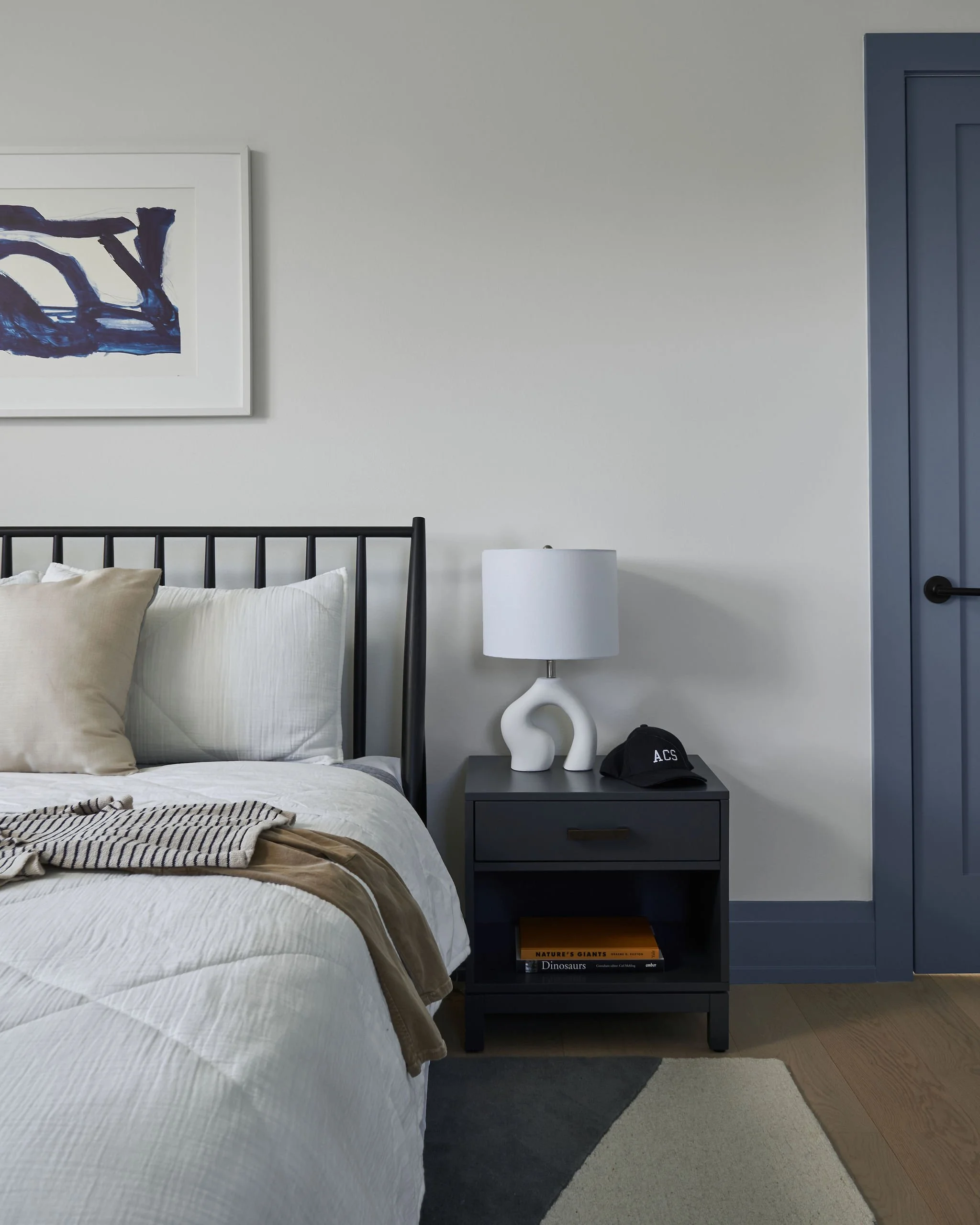 A bedroom with a bed, a nightstand with books, a table lamp, a decorative sculpture, a cap, and a framed abstract painting on the wall.