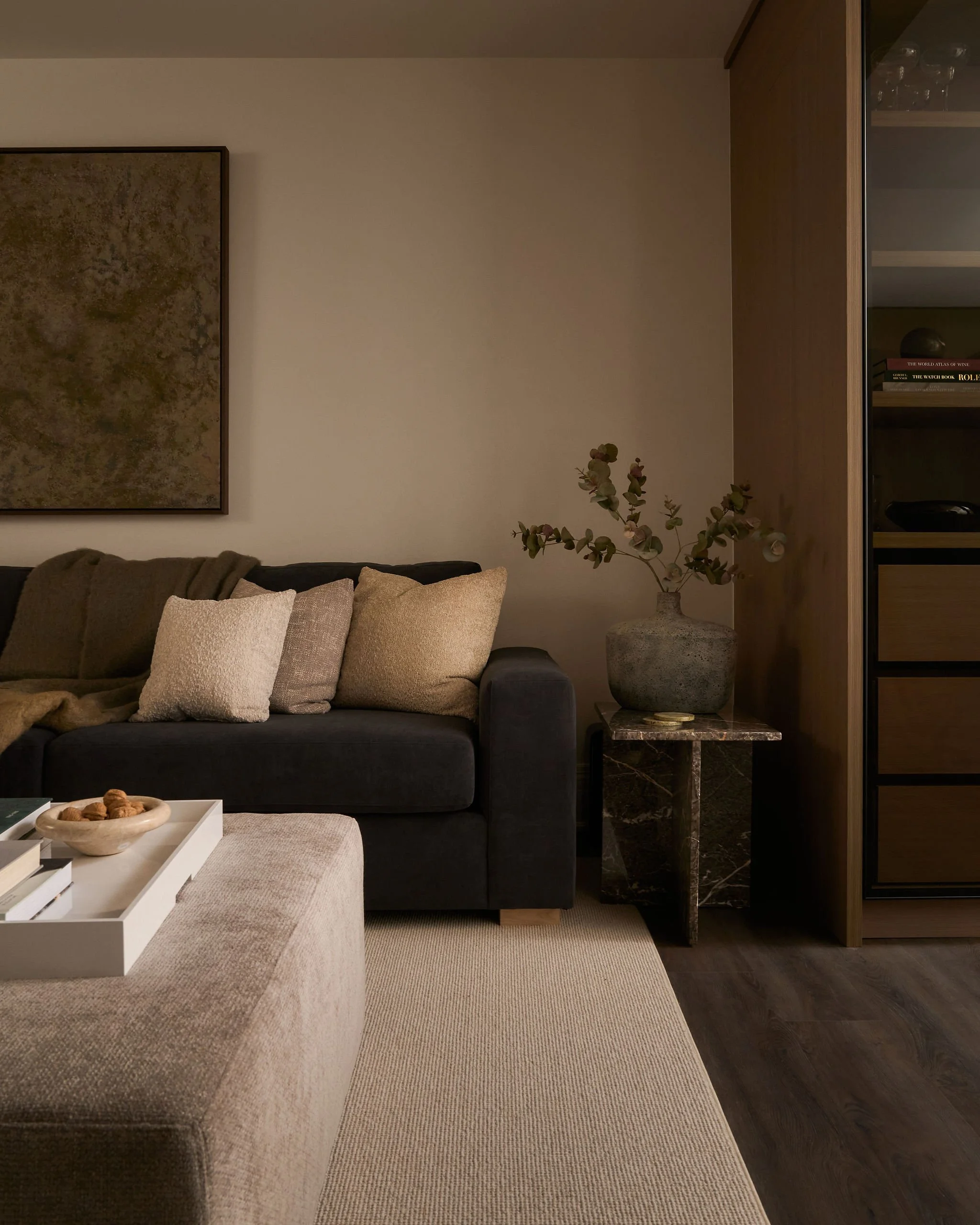 A living room with a dark grey sofa holding cream and beige pillows, a white coffee table with a bowl of mixed nuts, a beige and brown rug, a plant with green round leaves, and a wooden cabinet with glassware and books.
