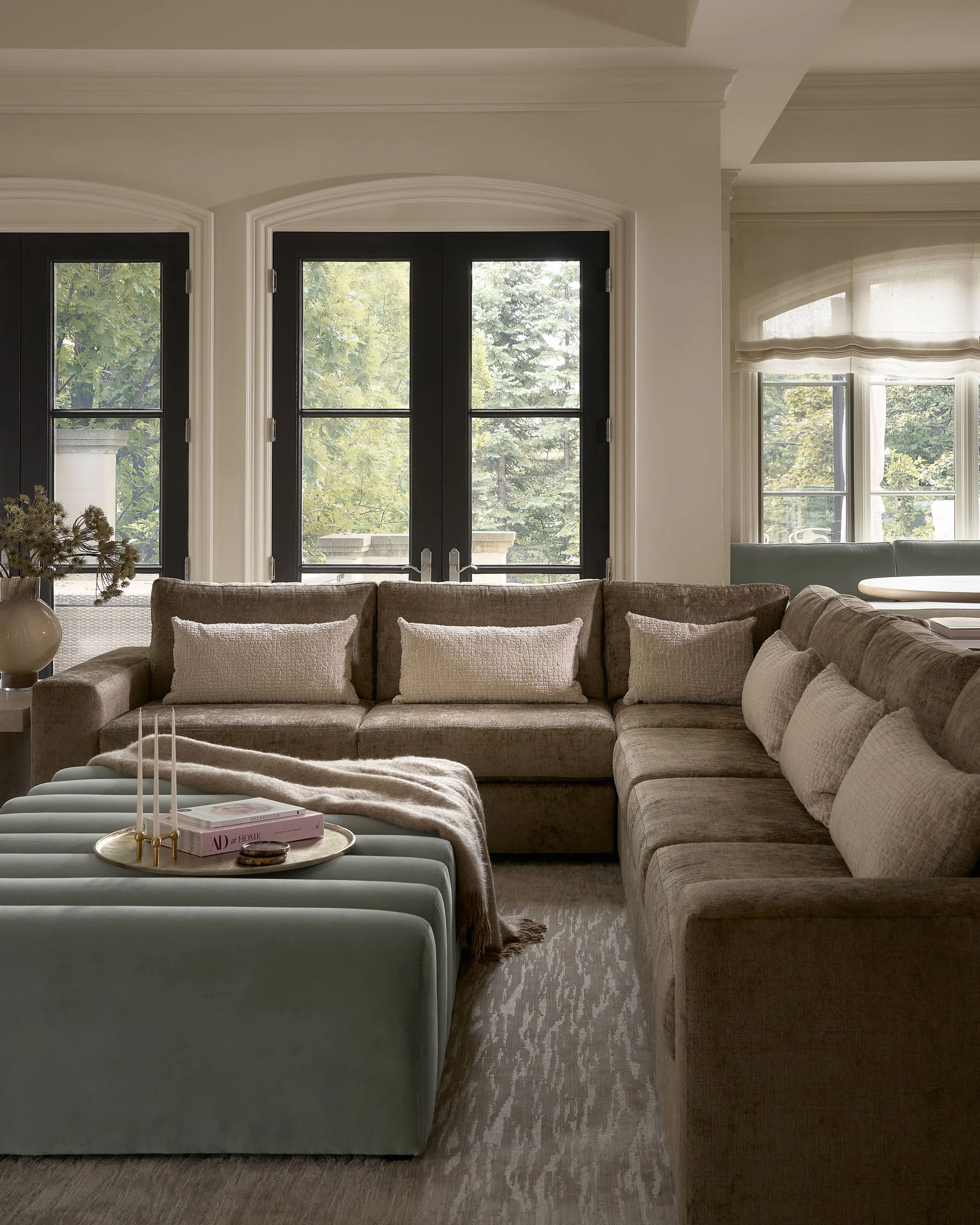 Living room with large beige sectional sofa, cream throw pillows, light rug, and a coffee table with candle holders, candles, and a pink magazine. Large window with white curtains, sunlight outside, and trees visible.