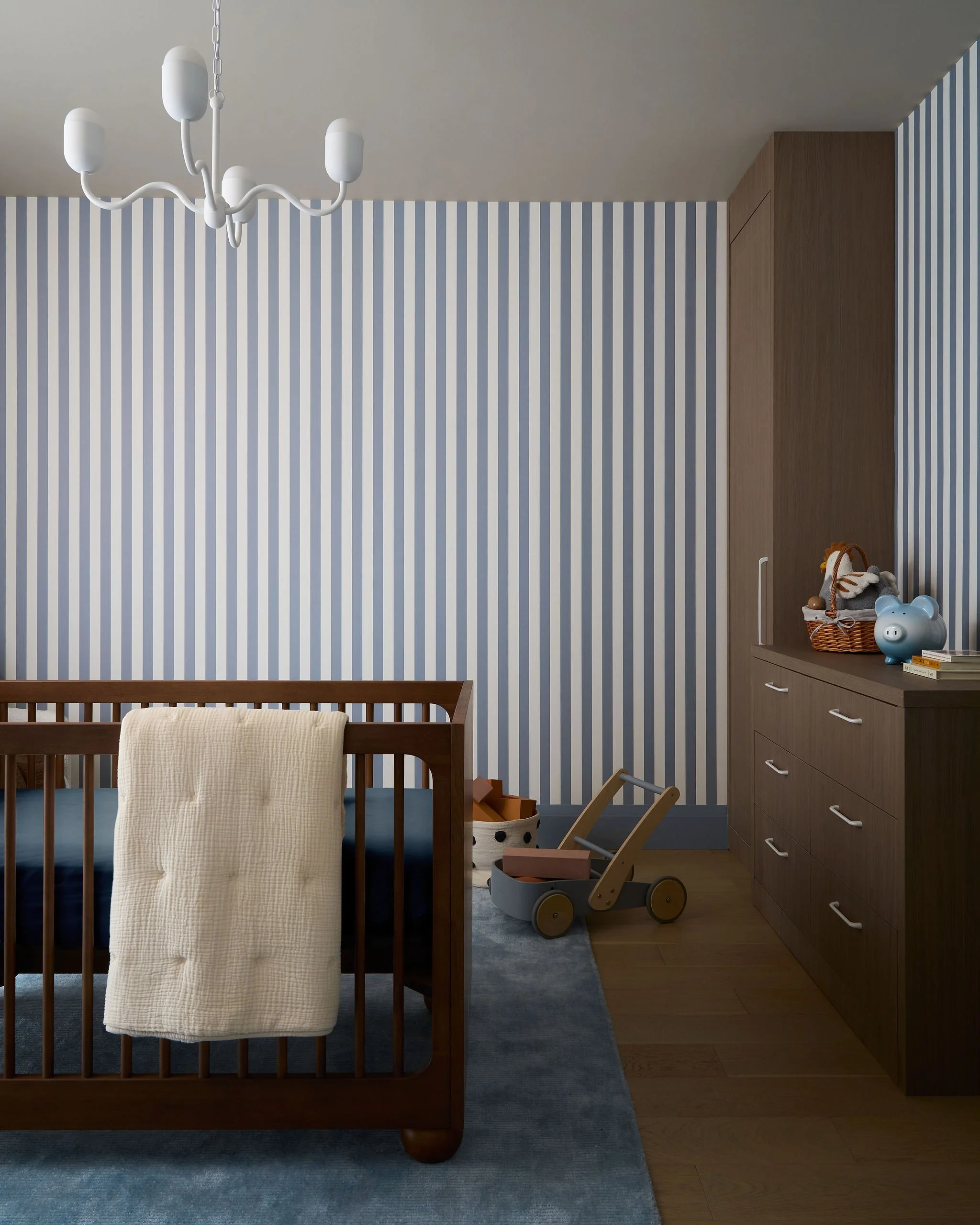 A child's bedroom with a wooden crib, a blue-gray rug, a beige blanket draped over the crib, a wooden toy cart, a wardrobe, and children’s toys including a blue piggy bank and a basket of stuffed animals.