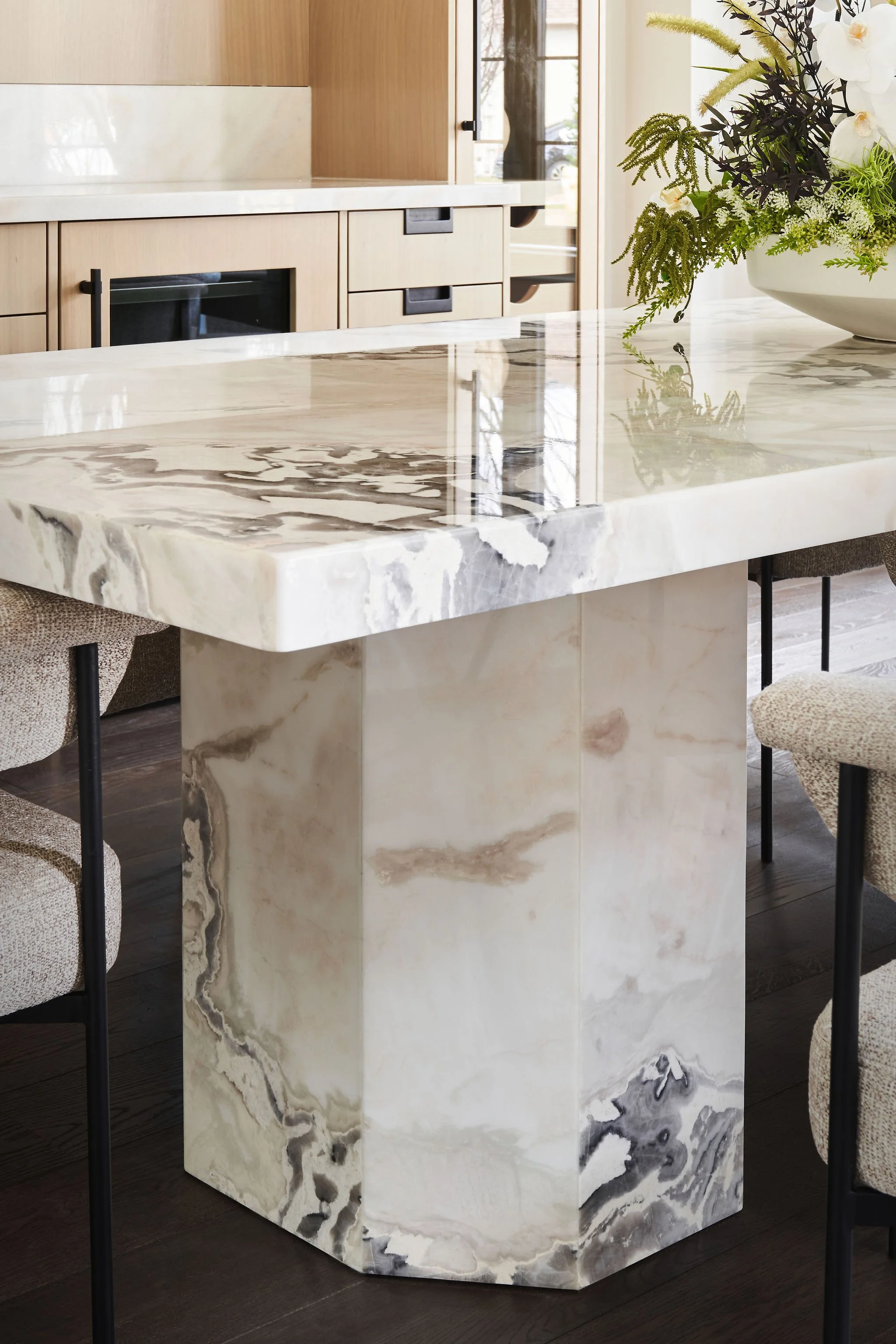 A marble island with a veined pattern in shades of beige, black, gray, and white in a modern kitchen with beige chairs and a floral arrangement.
