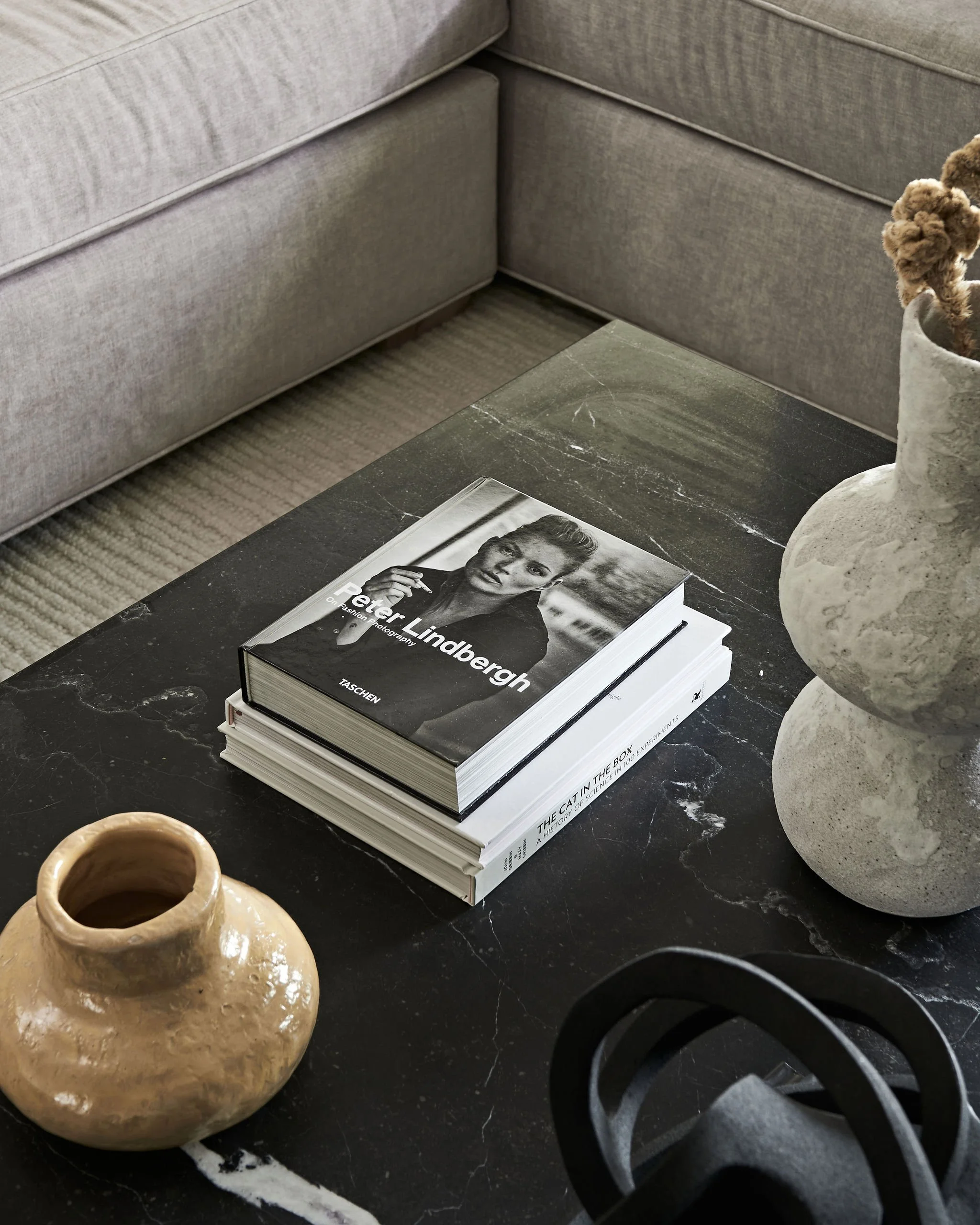 A table with a stack of books, a ceramic vase, a mug, and part of a black object in the foreground. The background shows a gray sectional sofa and a carpet.