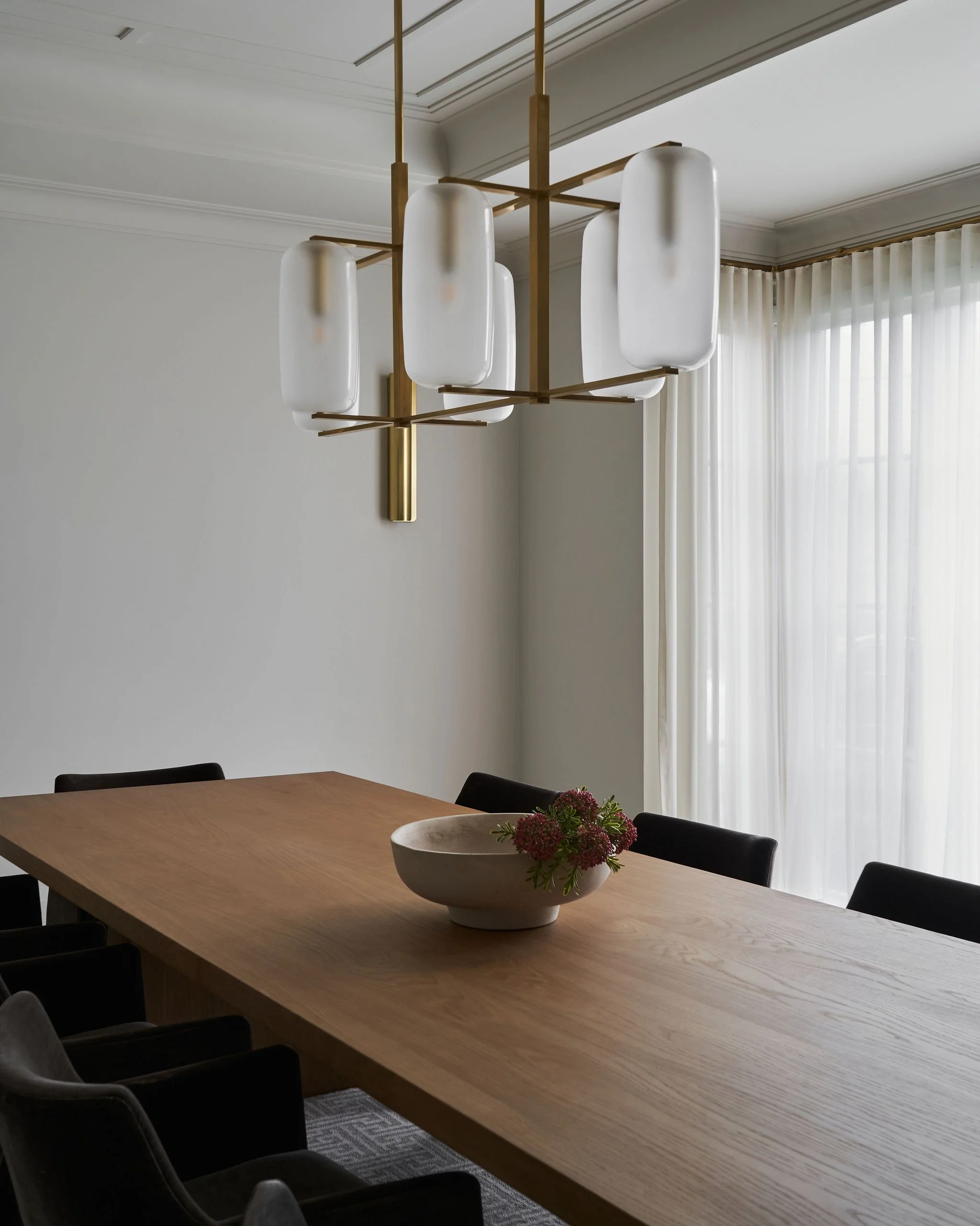 Modern dining room with a large wooden table, black chairs, and a minimalist white ceramic bowl with flowers. A contemporary chandelier with white shades and gold accents hangs above the table. Light filters through sheer white curtains in the backgr