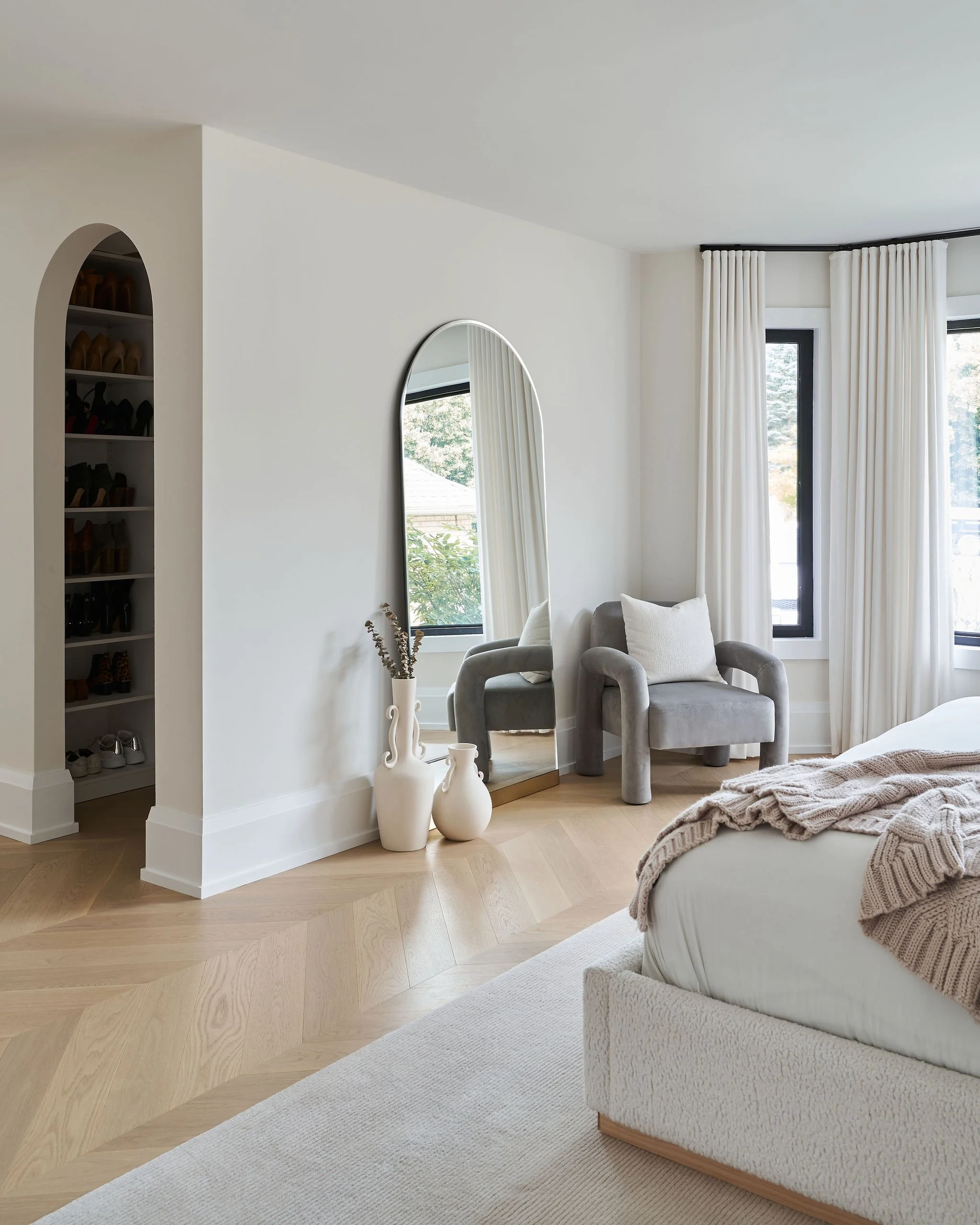 Bright and airy bedroom with light wood flooring, white walls, and large windows with white curtains. A cozy bed with a textured beige blanket is in the foreground. Two plush chairs with cushions sit near the window, and a tall mirror reflects part o