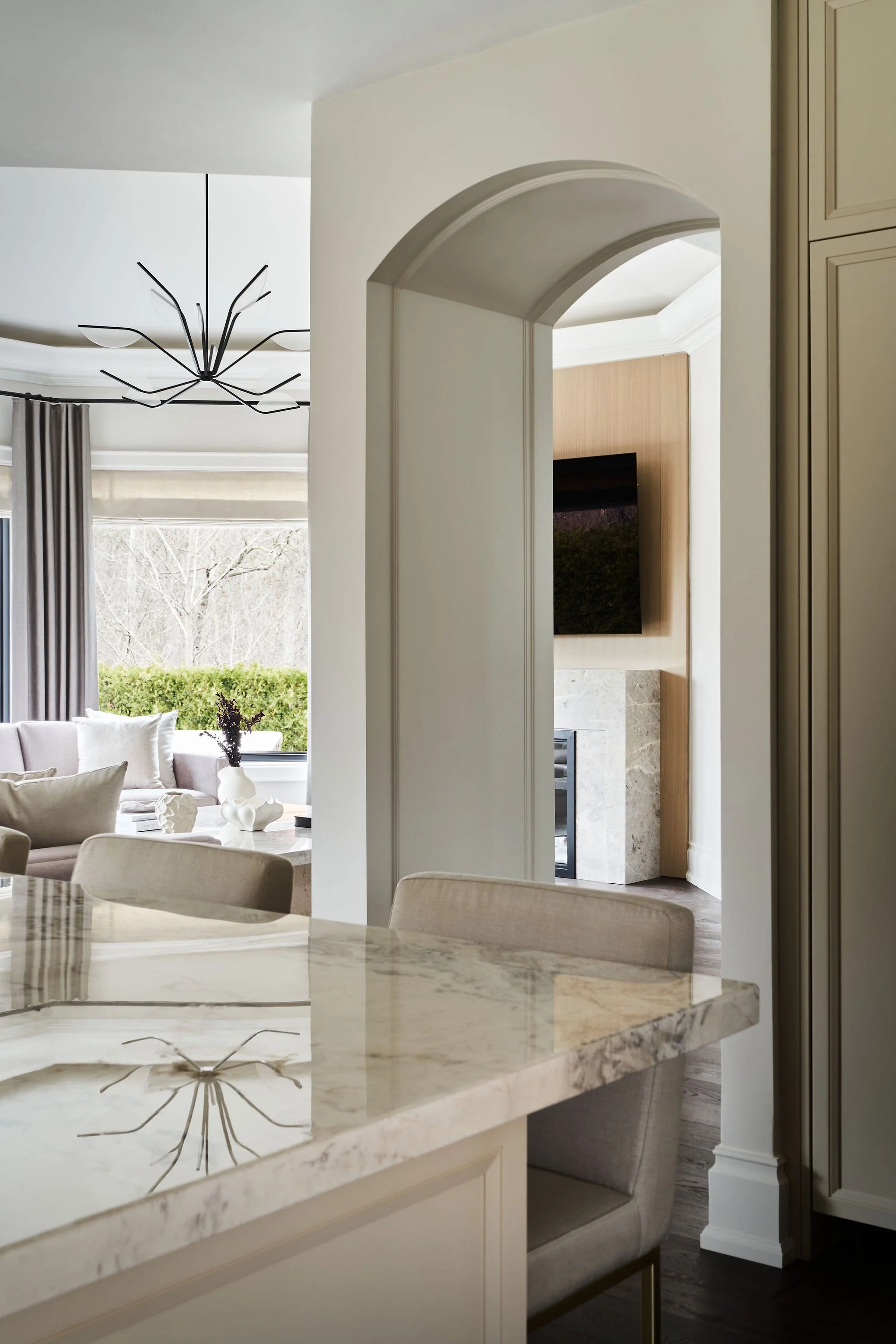 View of a modern living room and kitchen with a marble countertop, beige dining chairs, a beige sofa with white cushions, and a black branch-style chandelier. Large windows with gray curtains reveal an outdoor scene with trees and greenery.