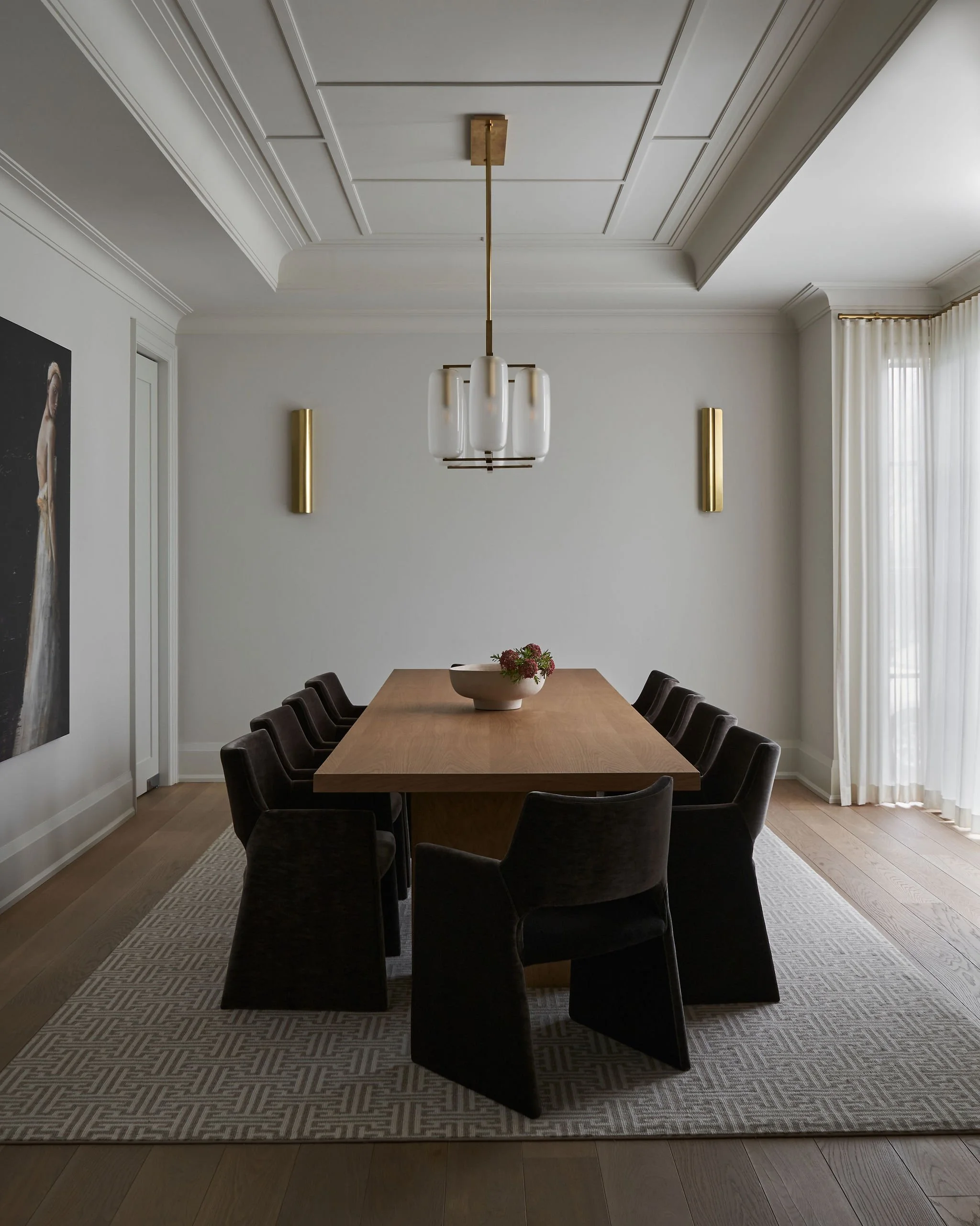 A modern dining room with a long wooden table, black upholstered chairs, a patterned rug, white walls, and a large window with white curtains. A vase with flowers is on the table, and a geometric pendant light hangs from the ceiling.