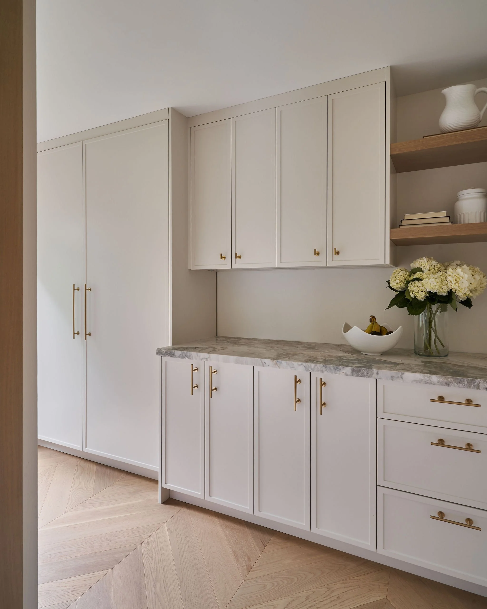White kitchen cabinets with gold handles, a marble countertop, a vase of white hydrangeas, a bowl of bananas, and two wooden floating shelves with decorative items.