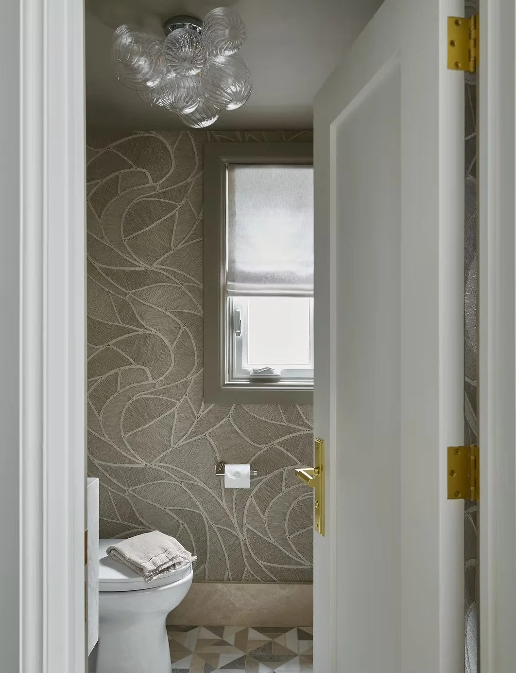 Small bathroom with a patterned beige wallpaper, a window with a frosted shade, and a toilet with a towel on top. The ceiling has a modern glass bubble chandelier, and the floor features geometric-patterned tiles.