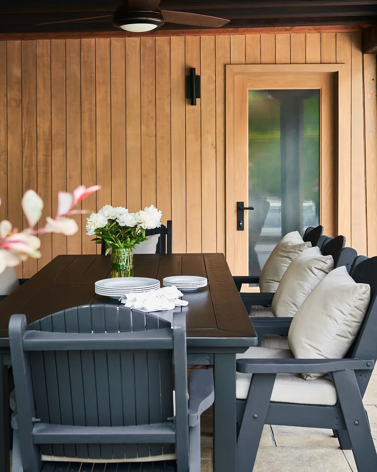 A dining area with a dark wooden table, beige cushioned chairs, and a gray chair in front. The table has a vase with white flowers and a stack of white plates with napkins. The background features a wooden paneled wall, a glass door, and a black wall