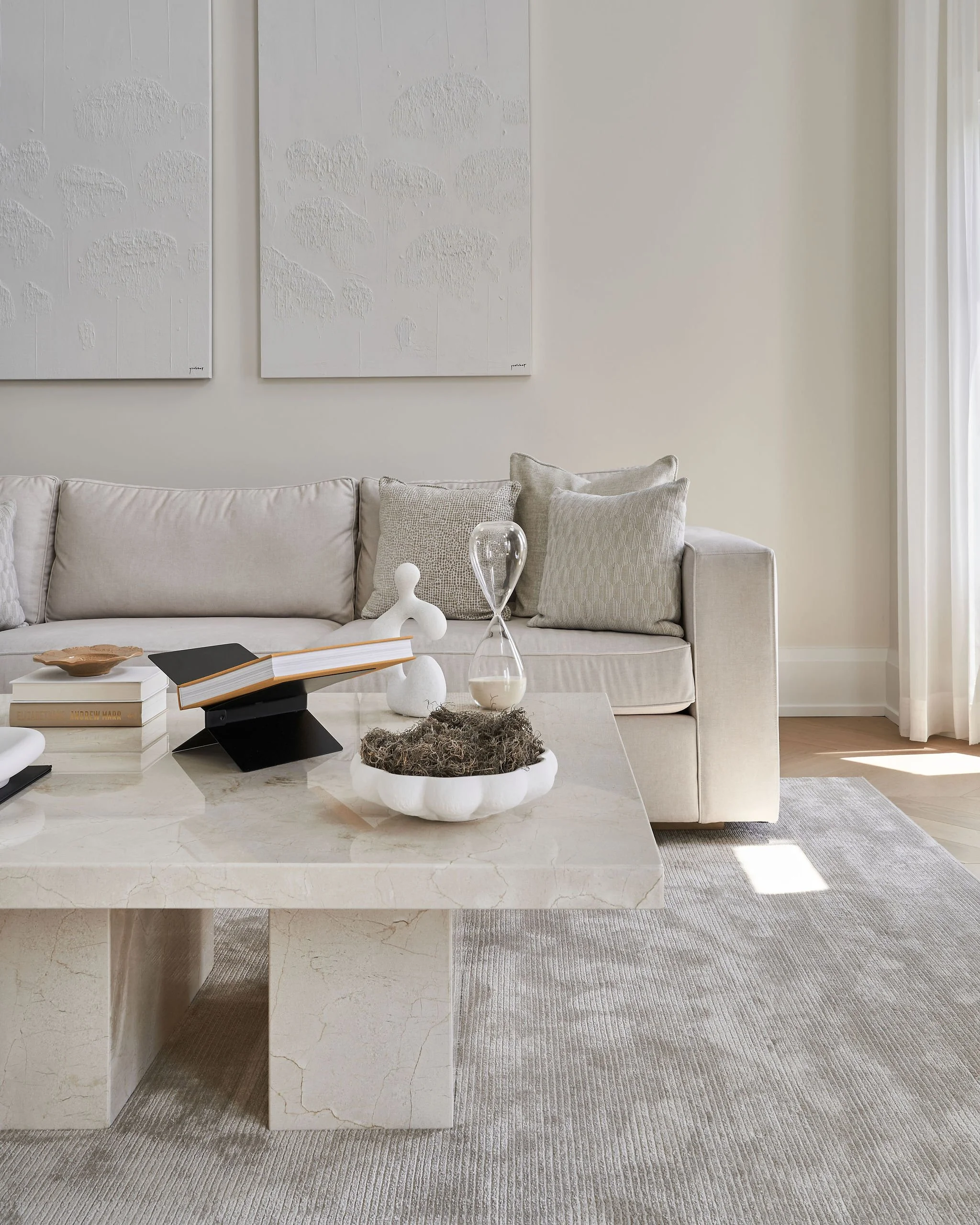 Living room with white sofa, decorative pillows, a coffee table with open books, a bowl, and sculptures, with large framed white textured artwork on the wall and a beige Rug.