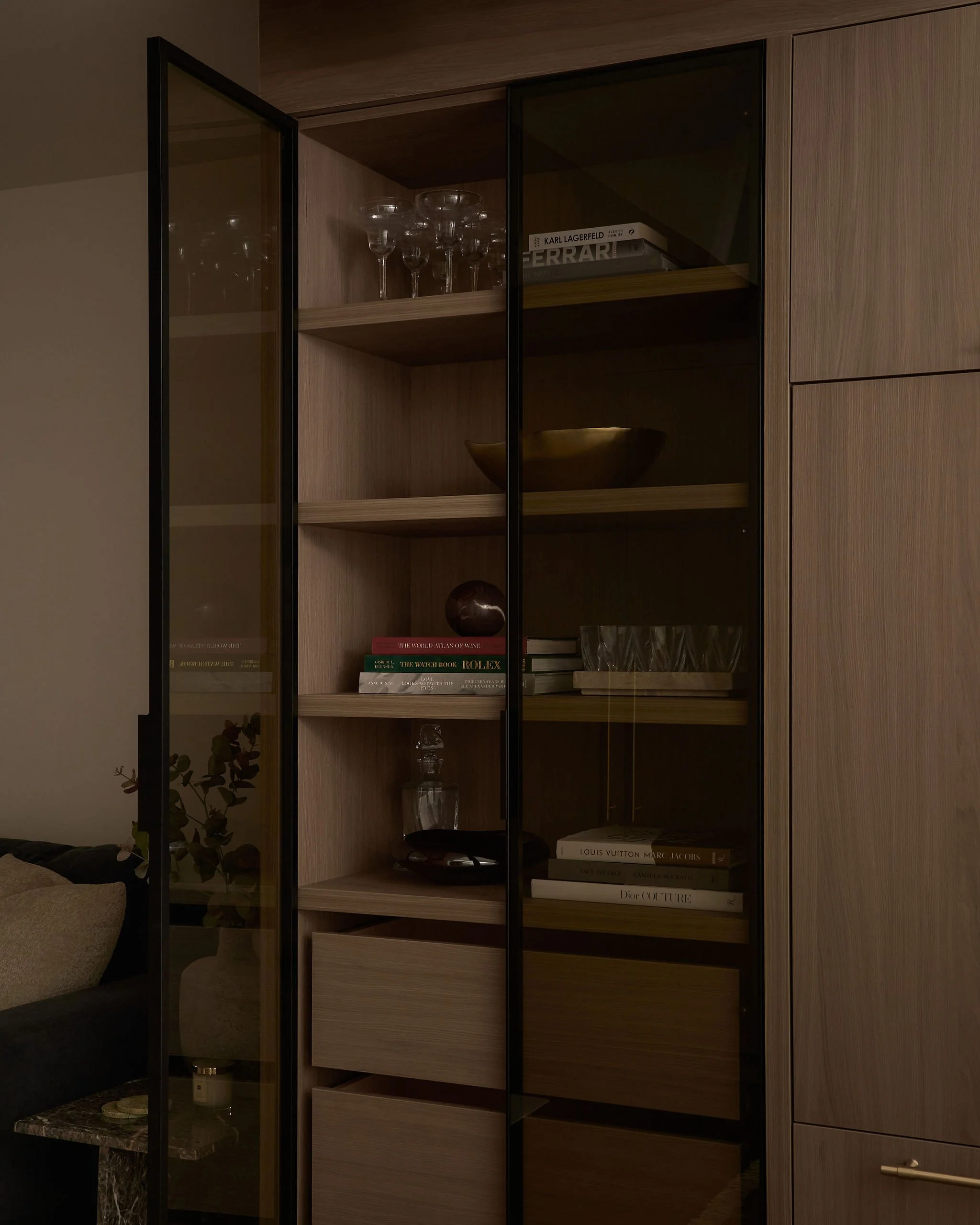 A wooden cabinet with glass doors containing wine glasses, books, a yellow bowl, a glass decanter, and decorative items, in a modern living space.