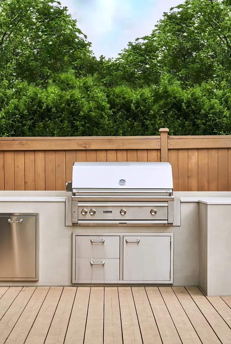 Outdoor kitchen area with a stainless steel grill, storage cabinets, and a wooden fence with lush green trees in the background.