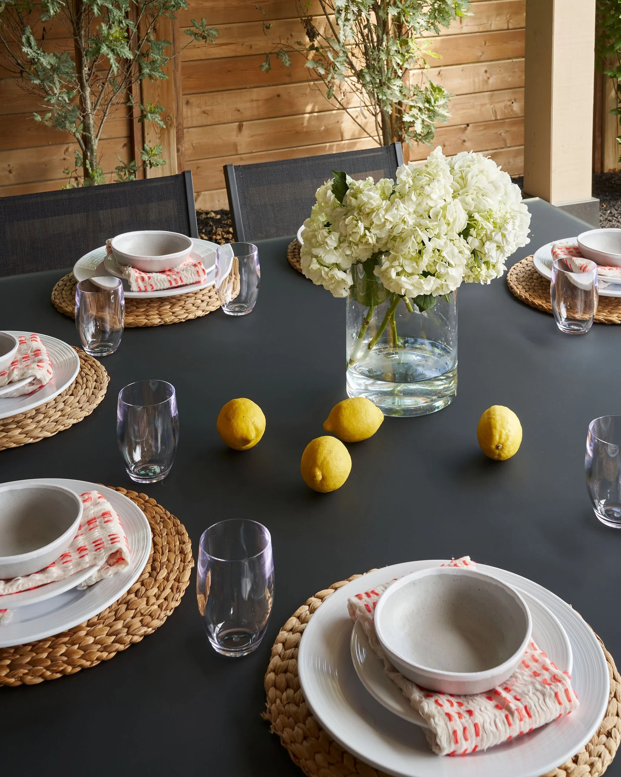Outdoor dining table with white hydrangea centerpiece, set with plates, bowls, glasses, and red-striped napkins on woven placemats, against a wooden fence and trees.