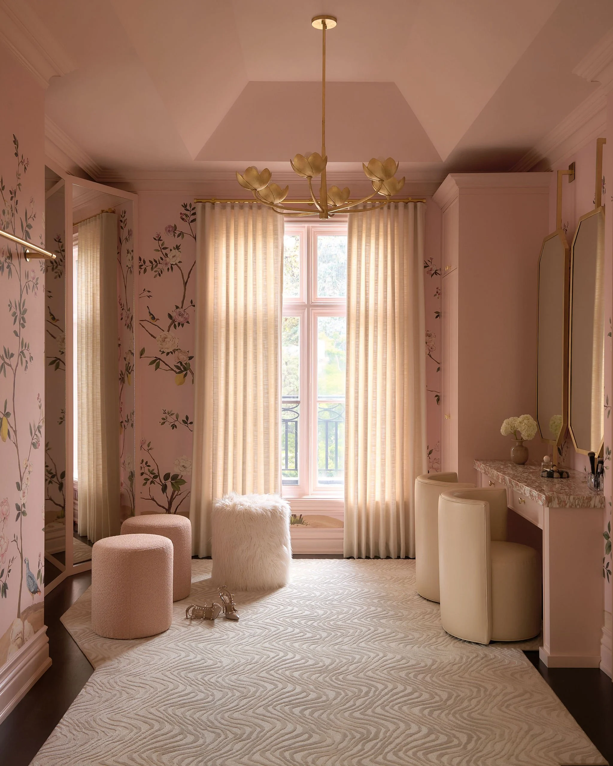 A feminine bedroom with soft pink walls and floral decals, featuring a large window with cream curtains, a chandelier with flower-shaped lights, a white furry stool, two pink upholstered stools, a vanity with a marble top, and a set of white bar stoo