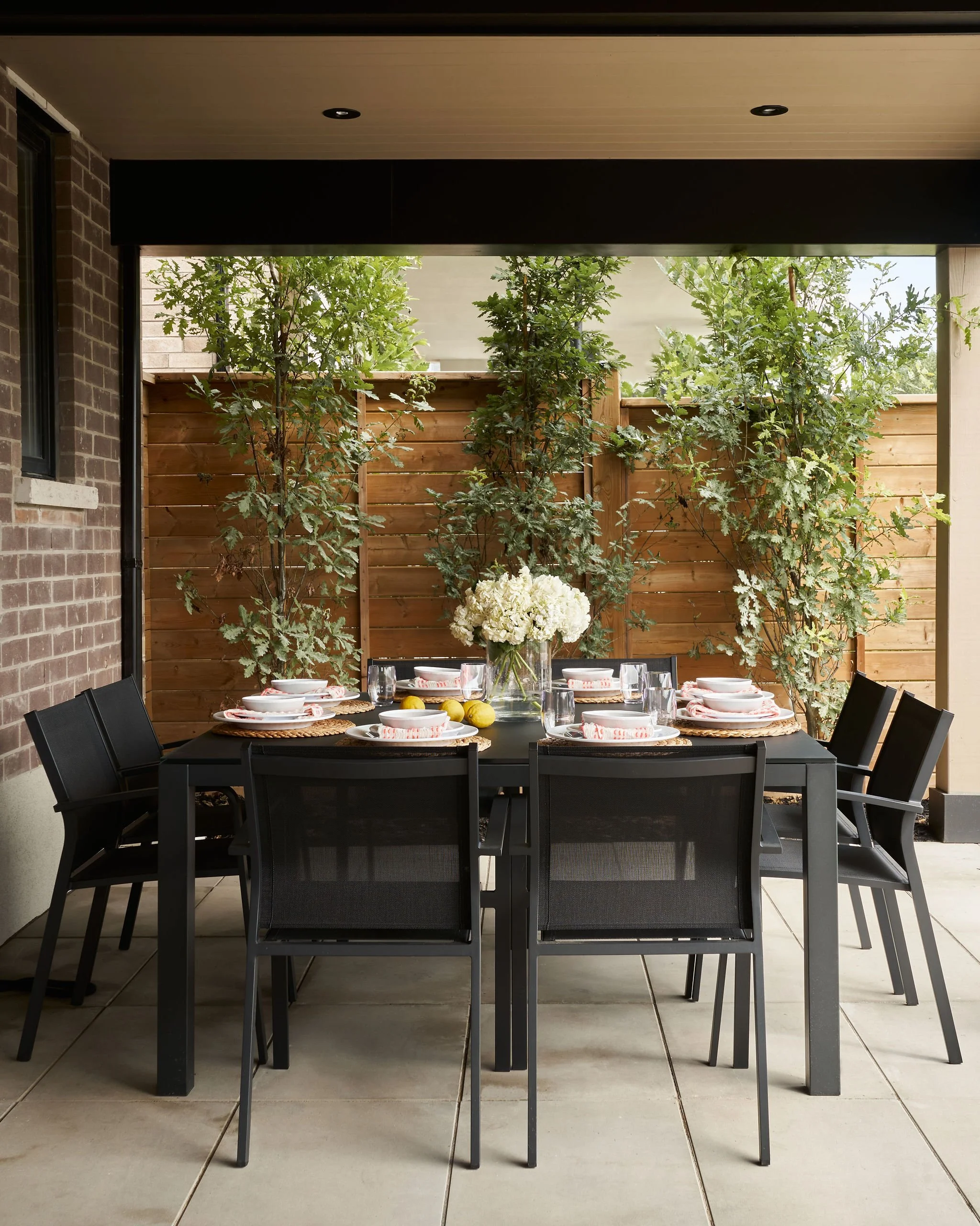 Outdoor dining area with black table set with plates, glasses, and napkins, surrounded by black chairs, featuring a floral centerpiece with white flowers, against a background of green trees, a wooden fence, brick wall, and ceiling with recessed ligh